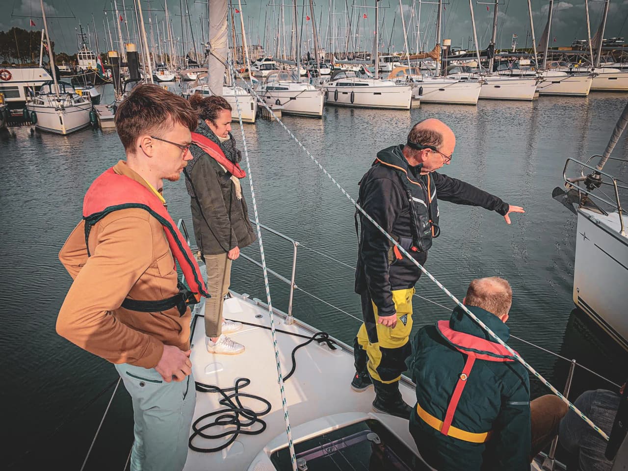 Vier mensen op een zeilboot in Zeeland, leren navigatiemanoeuvres onder een bewolkte hemel.