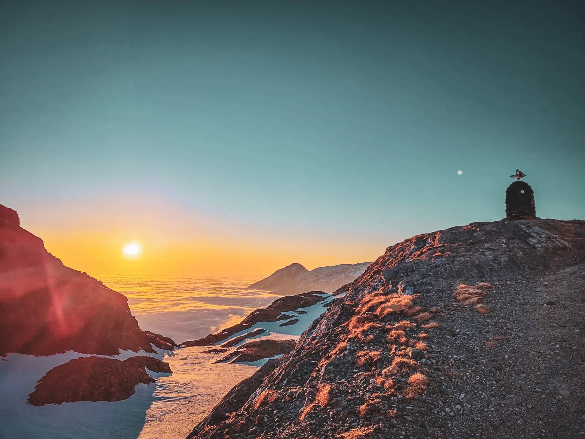 A mountain landscape at dawn, as the sun rises over the horizon. The sun's rays cast a golden light on the surrounding peaks, while a sea of clouds stretches across the horizon.