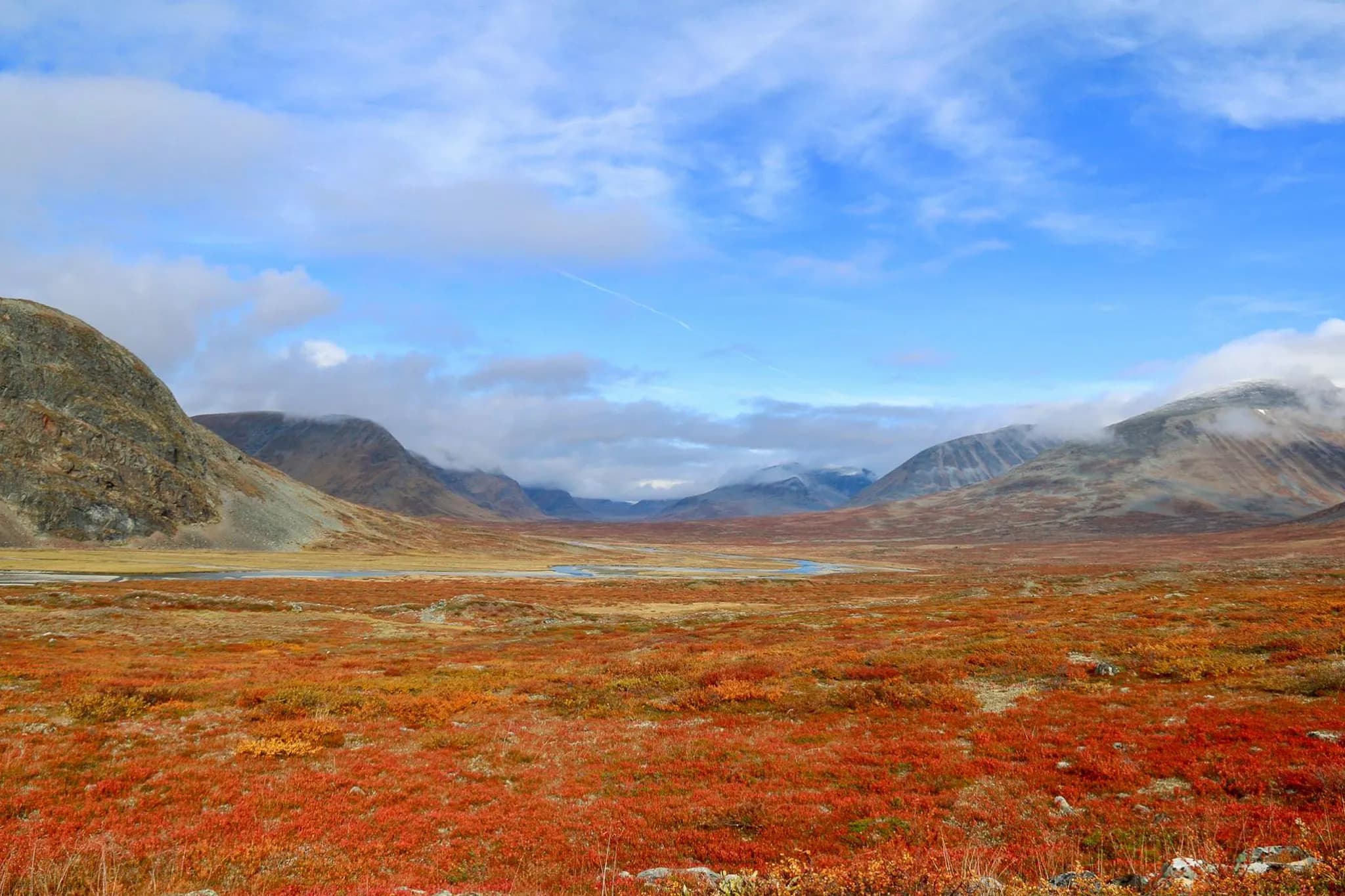 A vast, wild landscape with mountains in the background, partial clouds in a blue sky. The ground is covered with autumn-coloured vegetation, mainly in shades of red and orange, creating an atmosphere that is both warm and welcoming.