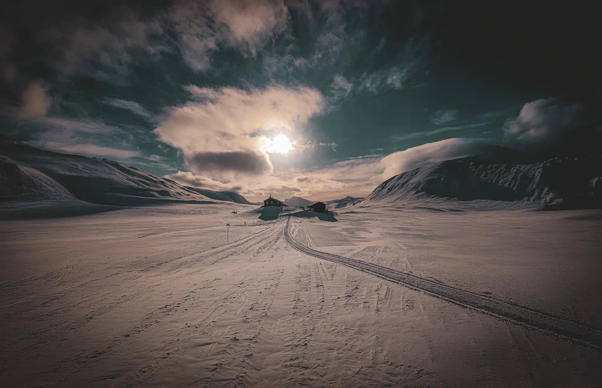 The snowy landscape of Swedish Lapland stretches as far as the eye can see, dominated by hills and mountains in the distance. Two buildings stand at centre stage, flanked by tracks left in the snow.