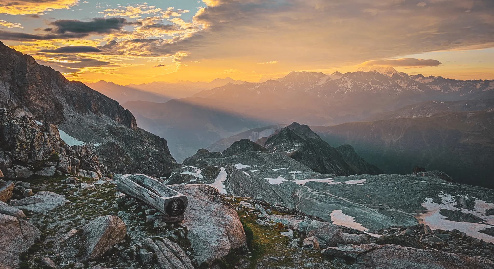 A breathtaking mountain landscape bathed in the golden light of sunset. The contours of the mountains are outlined in shades of orange and pink, while the shadows stretch across the surrounding valleys.