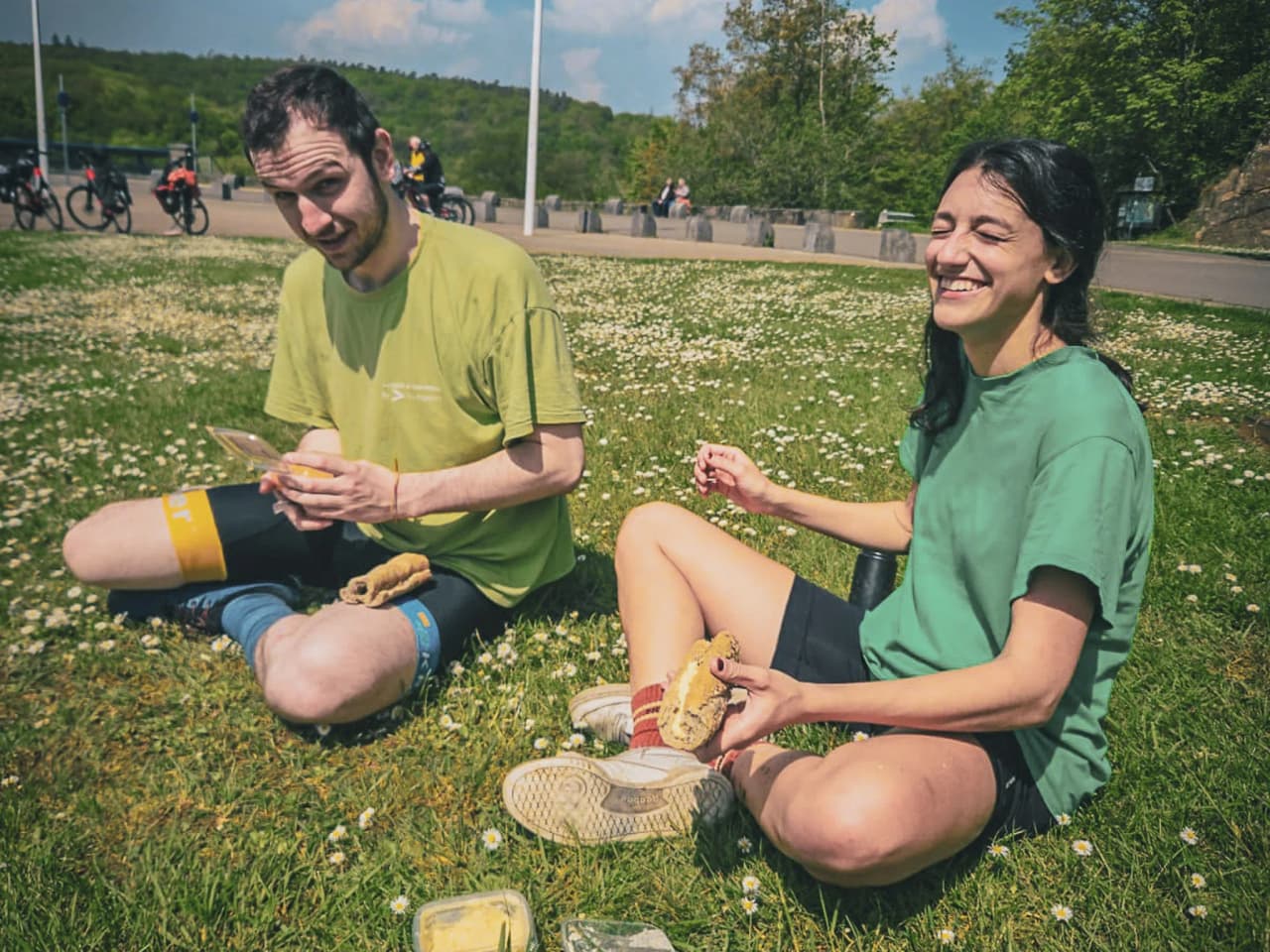 Deux aventuriers souriants dégustent un repas en pleine nature, entourés de fleurs et de verdure.