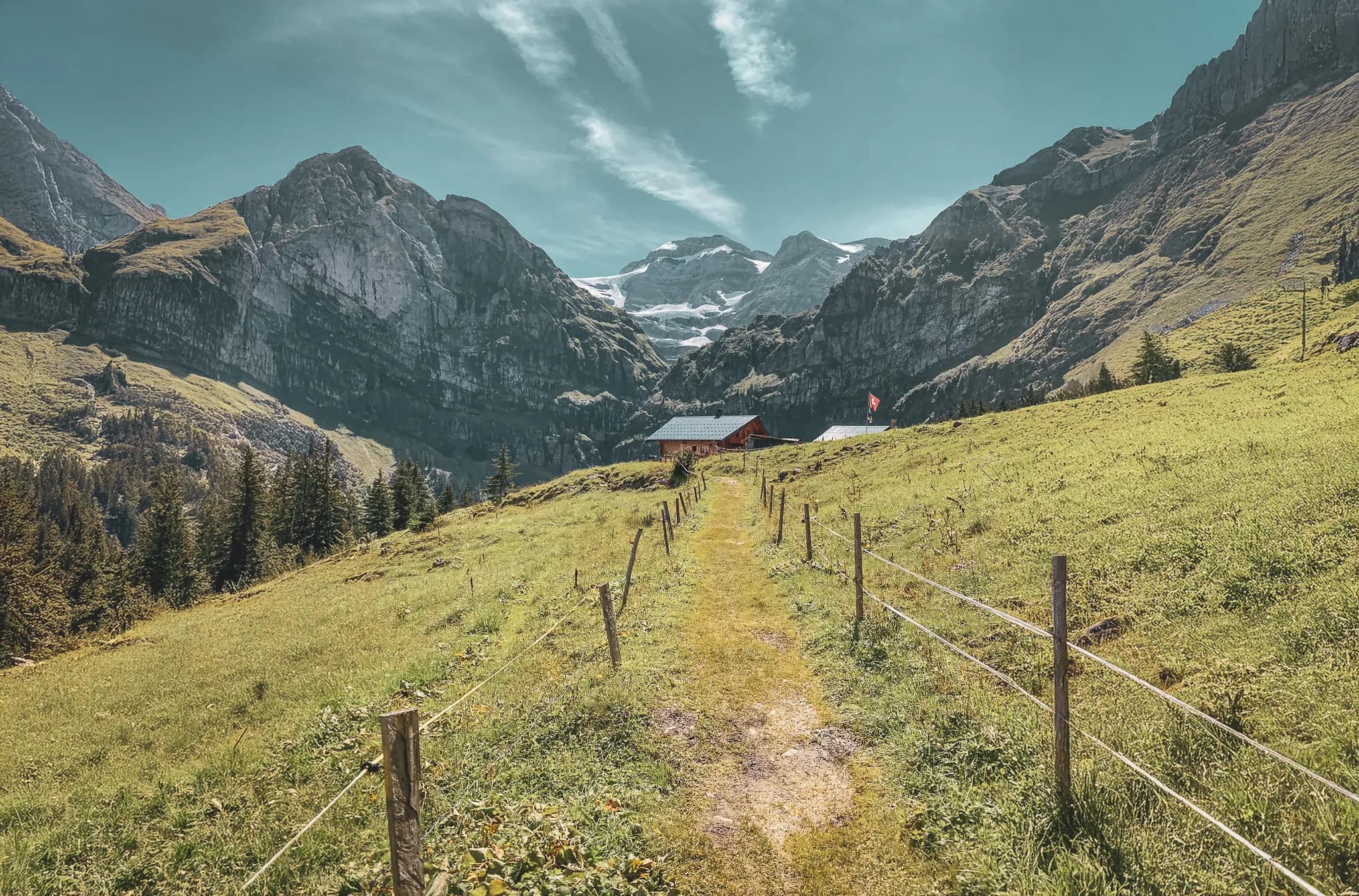 A leafy path winding through an alpine landscape, bordered by wooden fences, leads to a red wooden hut. In the background, majestic mountains covered in rock and snow.