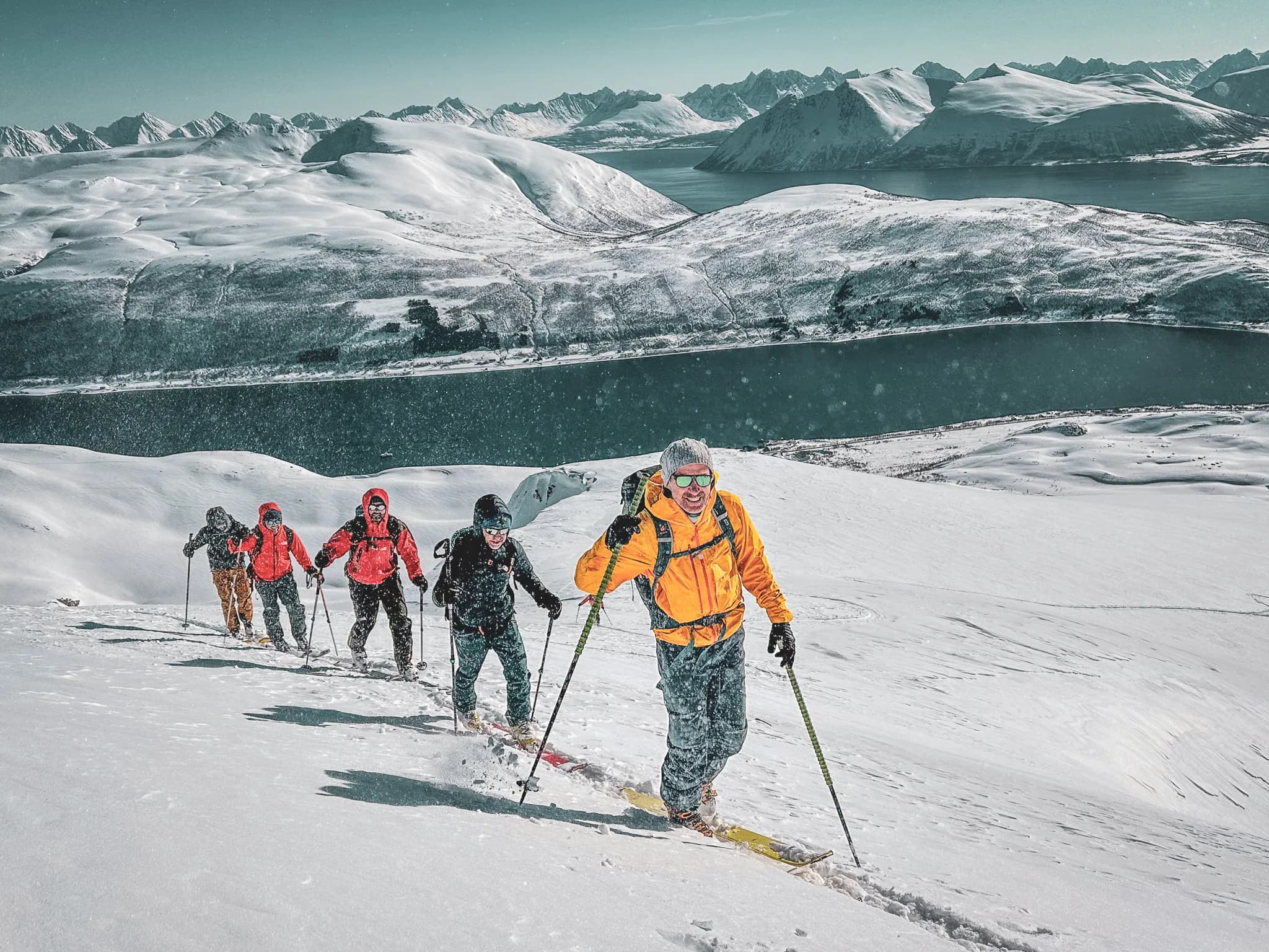 Un groupe de personnes en train de faire de la randonnée sur un terrain enneigé, avec des pics montagneux en arrière-plan. Les membres du groupe portent des vêtements de ski colorés, principalement des vestes rouges et une veste orange. Ils