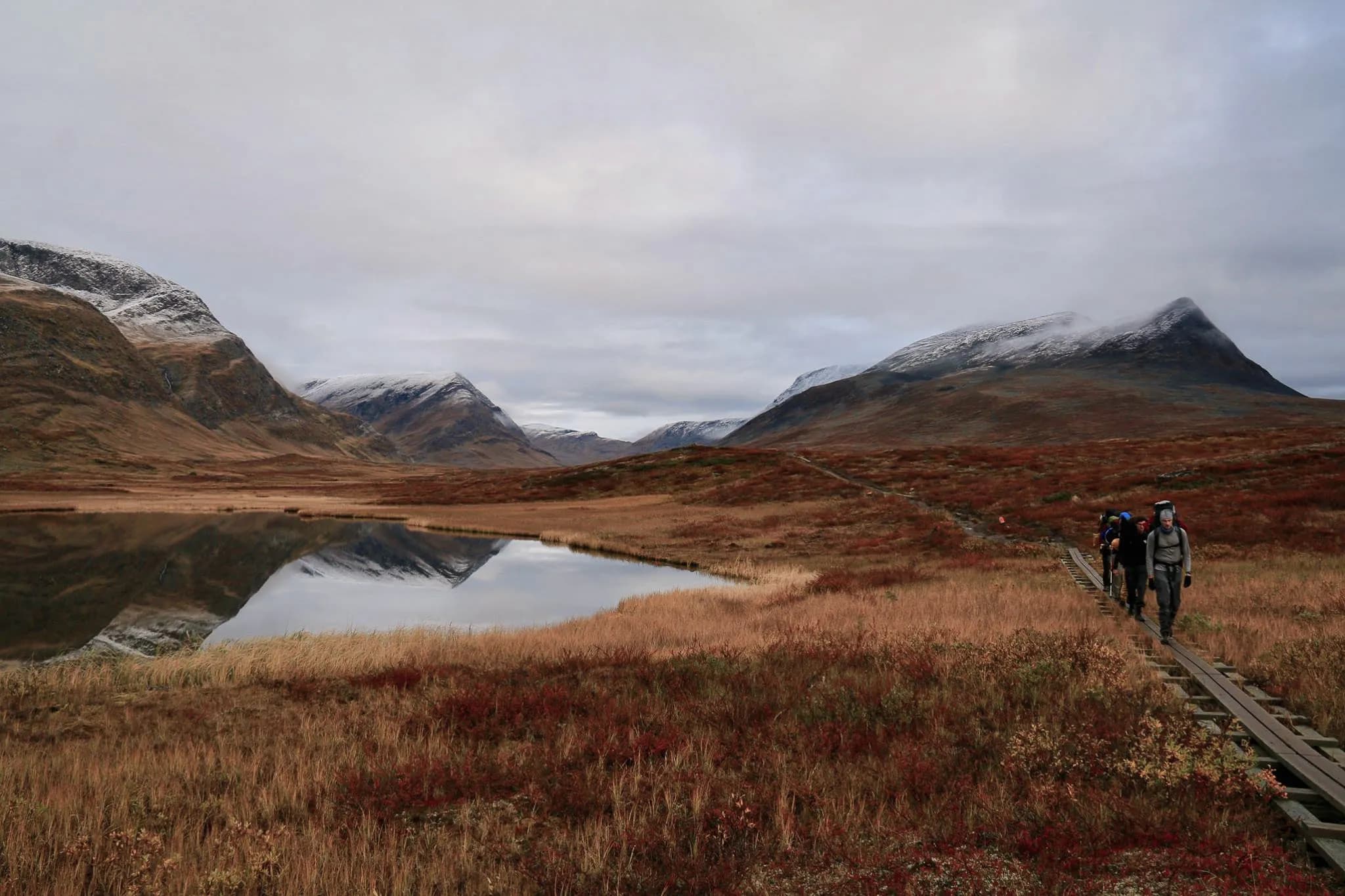 A mountainous landscape with snow-covered hills and gentle slopes. A calm lake reflects the mountains and the cloudy sky. In the lower part of the image, a wooden pathway is