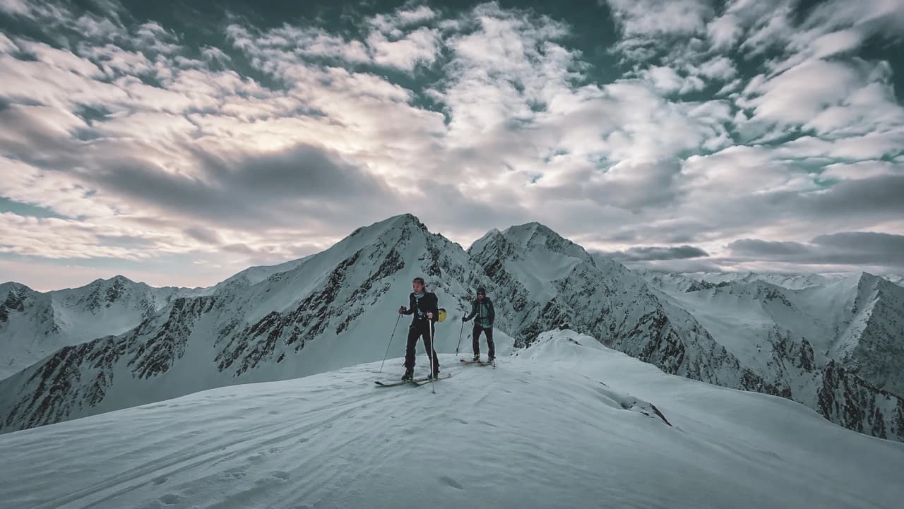 Two skiers make their way across a snow-covered peak, surrounded by majestic mountains beneath a cloudy sky.