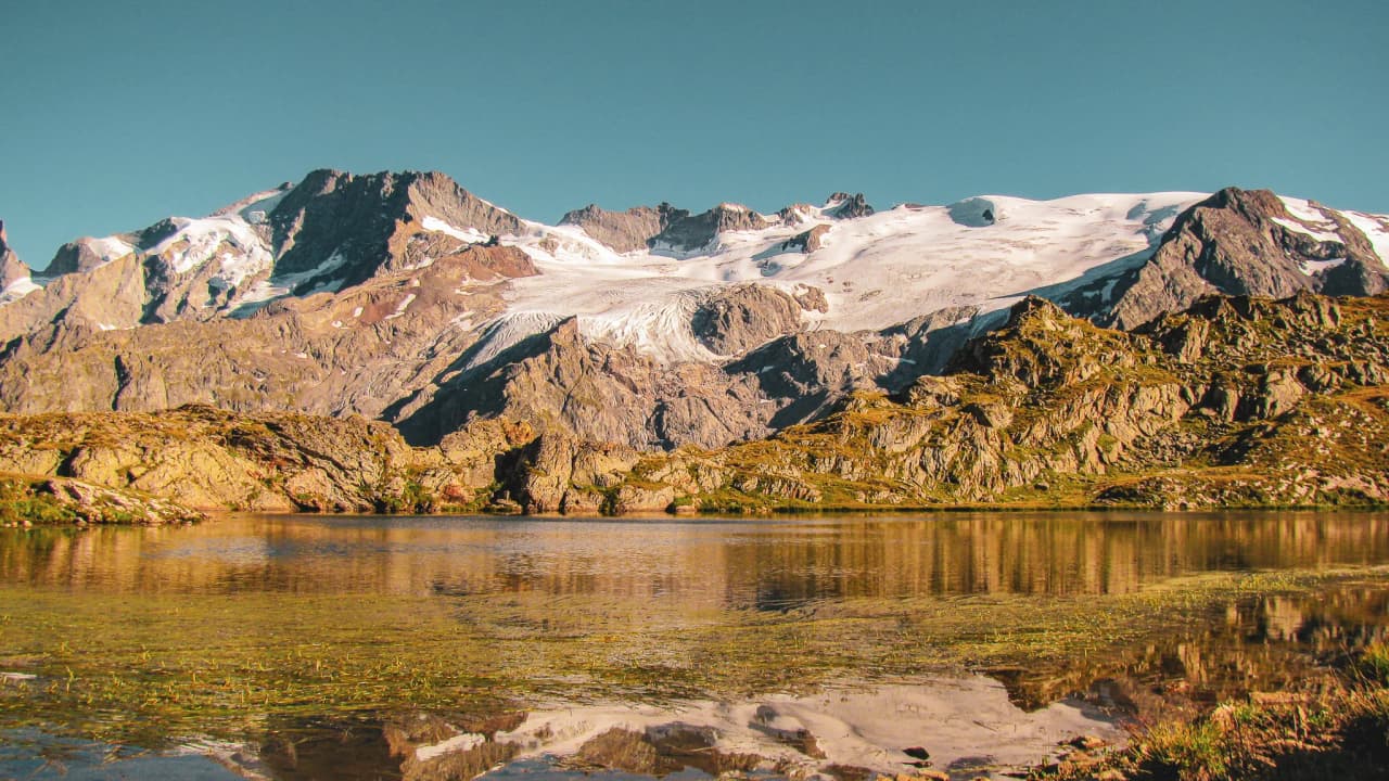 A peaceful lake surrounded by majestic mountains and glistening glaciers under a blue sky.