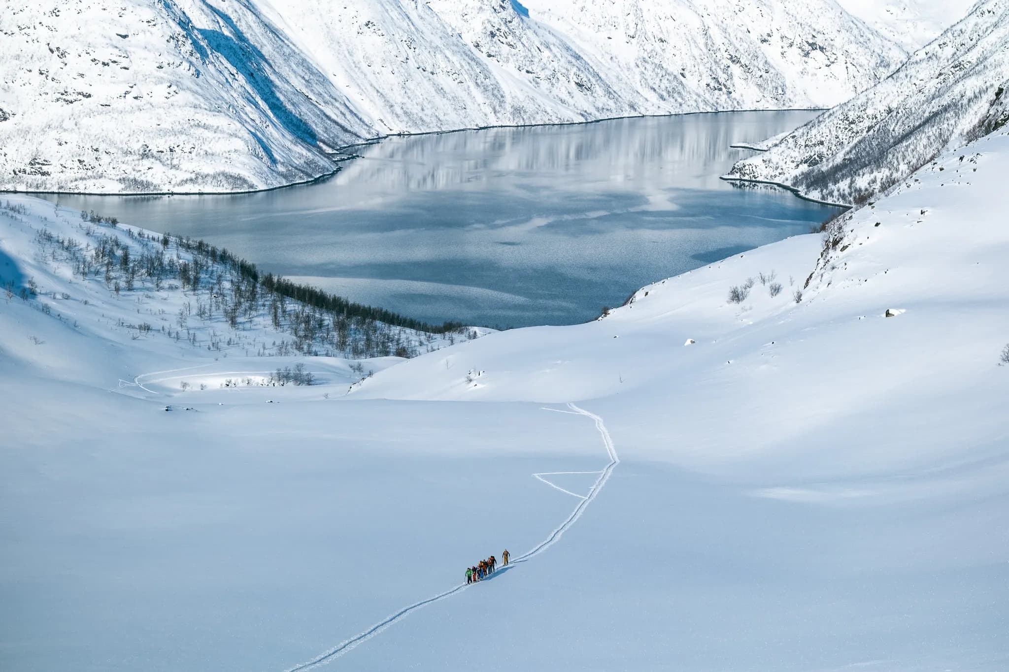 A snowy landscape with majestic mountains in the background and a tranquil lake reflecting the sky. In the foreground, a group of people are walking on the virgin snow, leaving tracks behind them. The scene is reminiscent of a