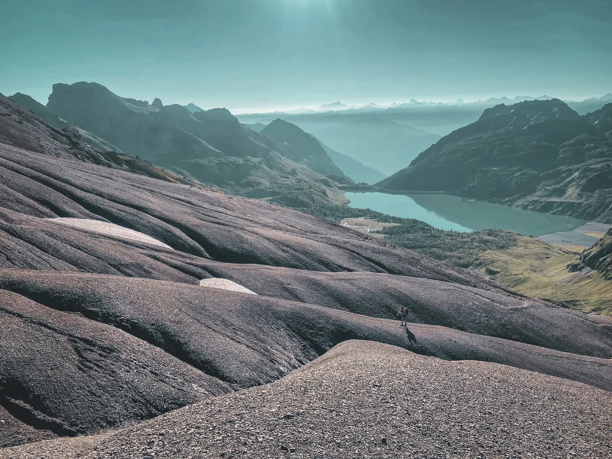 A spectacular mountain landscape, with undulating rock formations covered in dark gravel. In the background, majestic peaks loom, bathed in soft light. To the right, a lake
