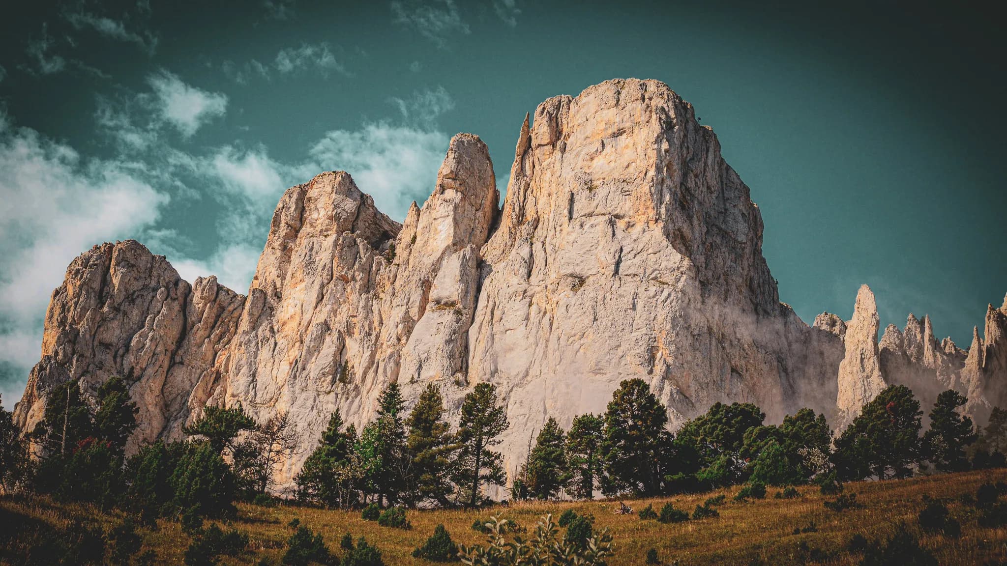 Majestic rock formations rise against a cloudy sky, interspersed with sunny spells. In the foreground, a forest of lush green coniferous trees contrasts with the craggy rocks.