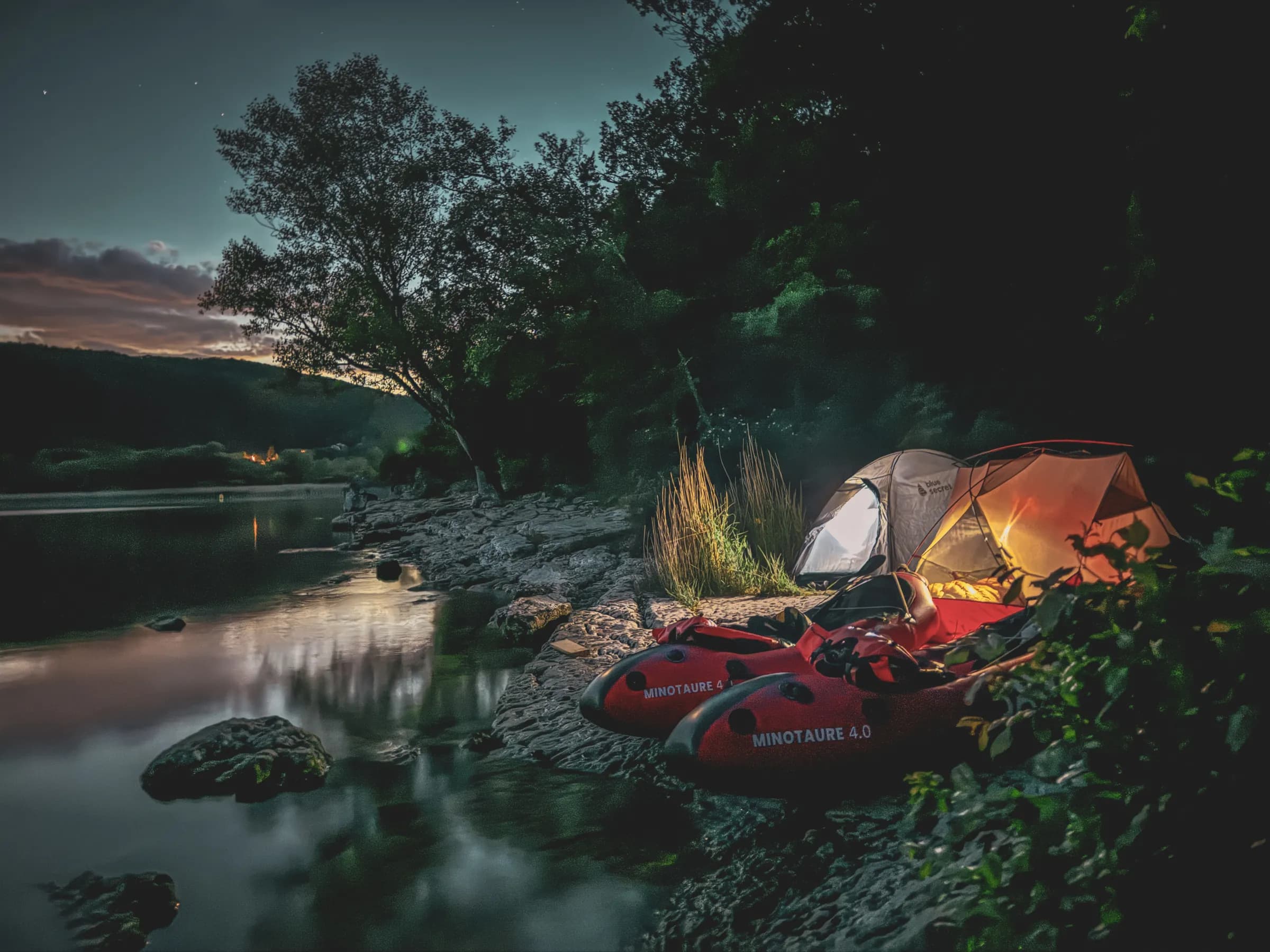 A night-time riverside camping landscape, with illuminated tents contrasting with the surrounding darkness. Two red kayaks are positioned nearby, resting on a bed of stones. The soft light