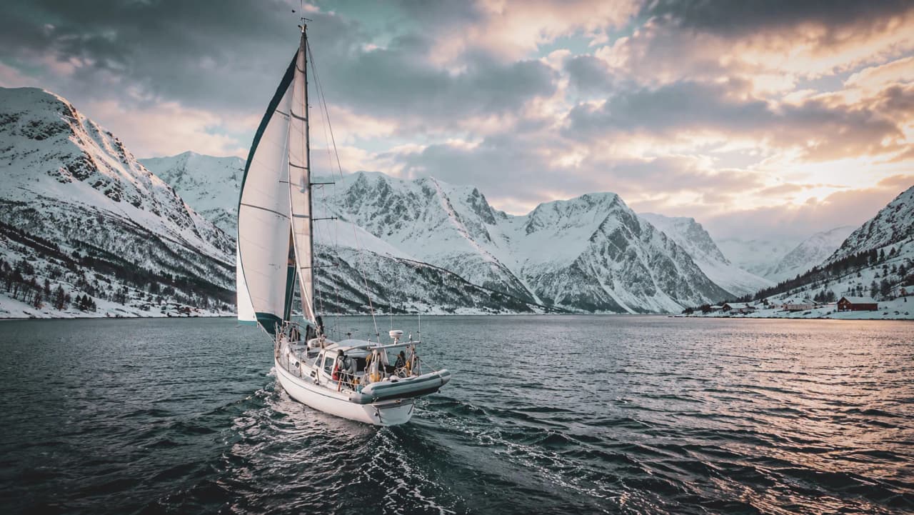 A sailing boat sailing through a snow-covered fjord, surrounded by majestic mountains and a bright sky.