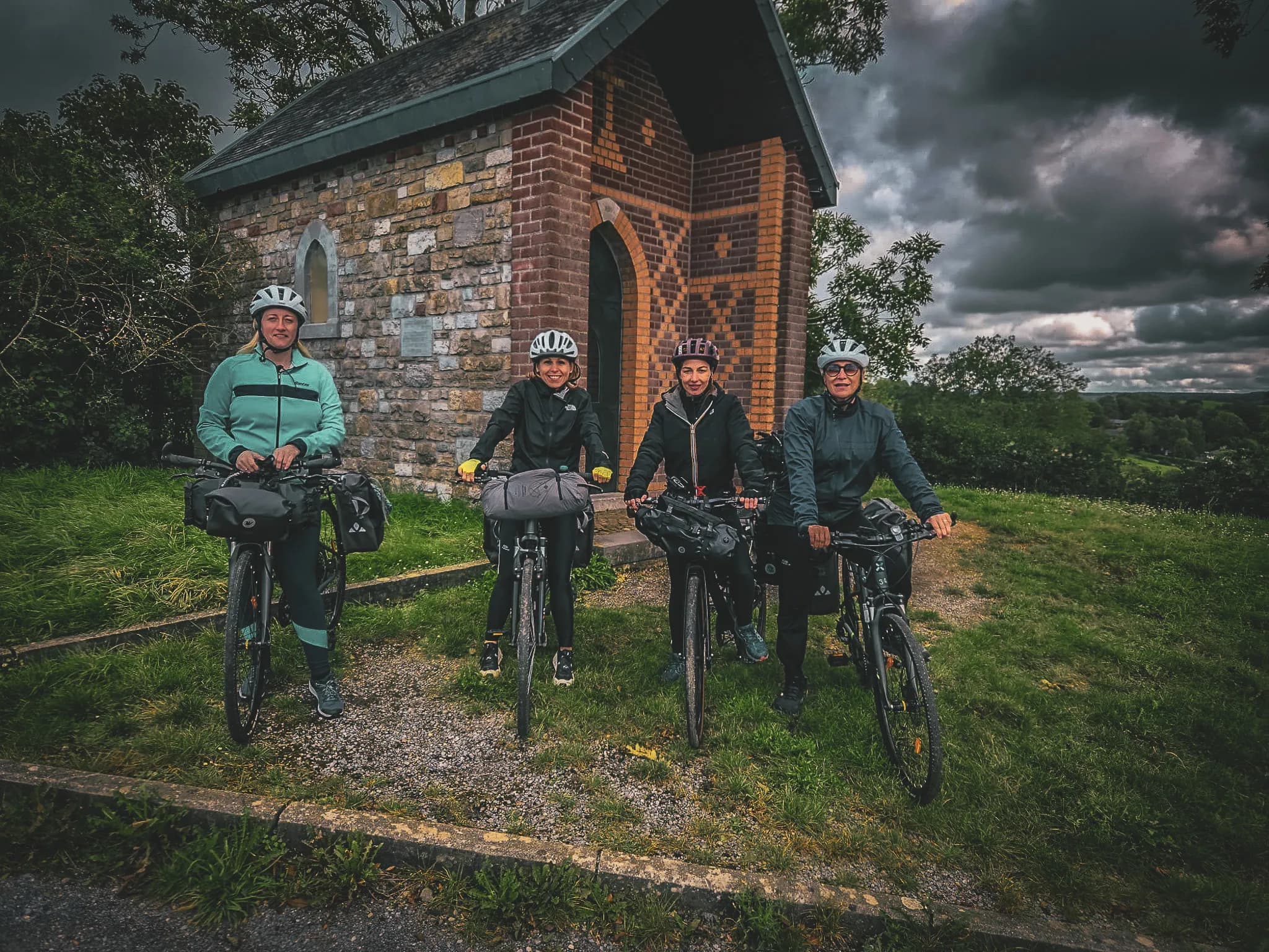Four cyclists pose in front of a small stone building, probably a chapel, surrounded by lush vegetation. They are wearing helmets and sports clothing suitable for cycling, with trekking bags strapped to their backs.