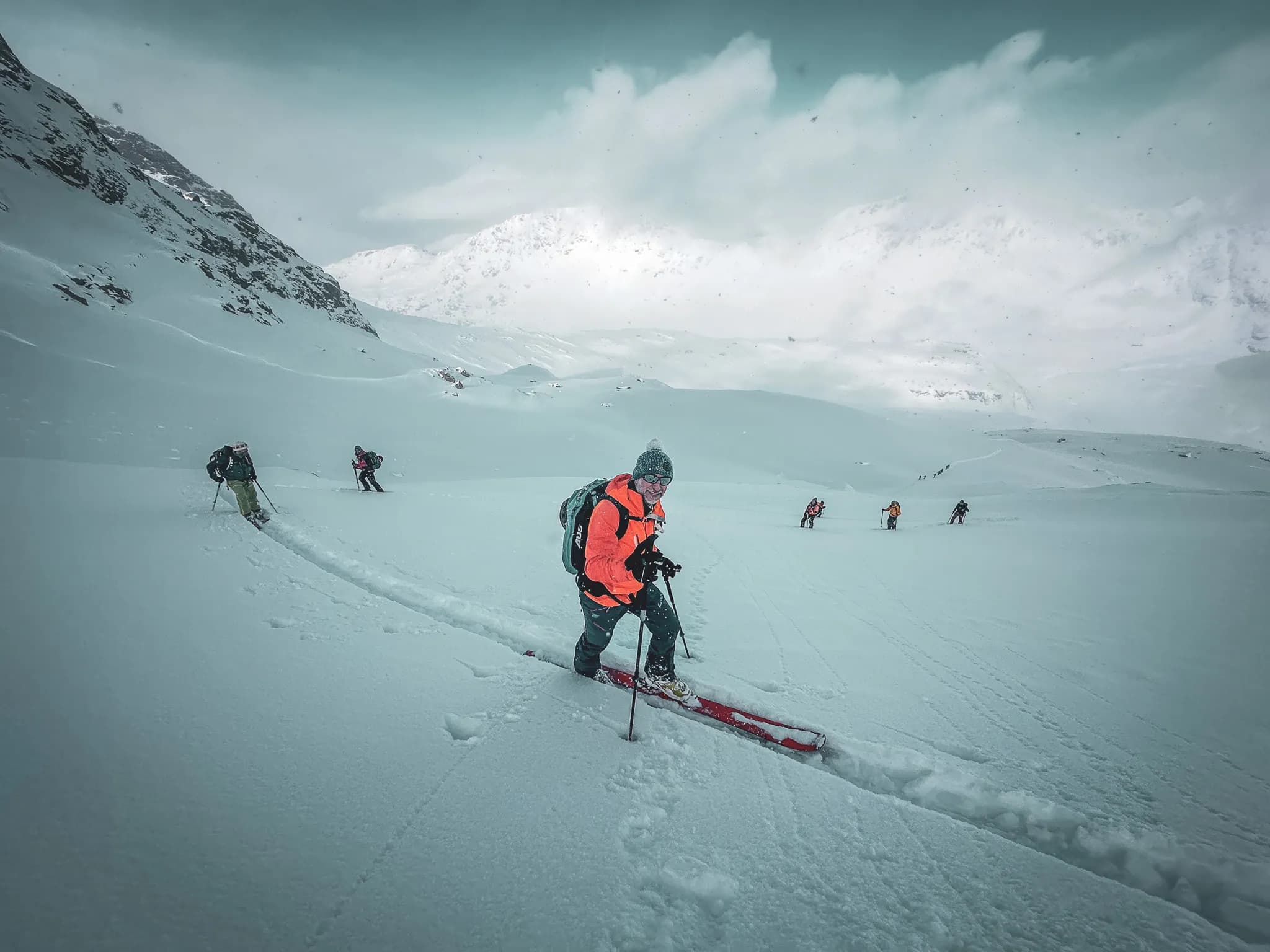 Un groupe de skieurs progresse sur une pente enneigée dans un paysage montagneux. L'un des skieurs, portant une veste orange et un bonnet, semble sourire tout en utilisant des skis et des bâtons pour avancer. En arrière-plan