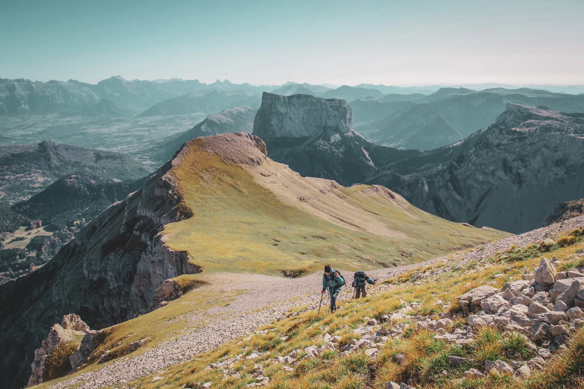 A spectacular mountain landscape with hikers walking along a steep path. The rock and green grass form a striking contrast, while the background reveals a mountain range.