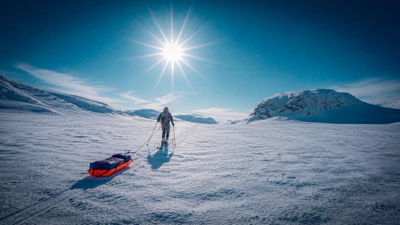Een avonturier die een pulka voorttrekt over een uitgestrekte besneeuwde vlakte, onder een stralend blauwe hemel.