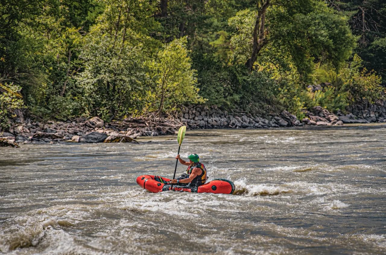Een avonturier in een packraft peddelt over een woeste rivier, omringd door groen. Ontsnapping en avontuur!