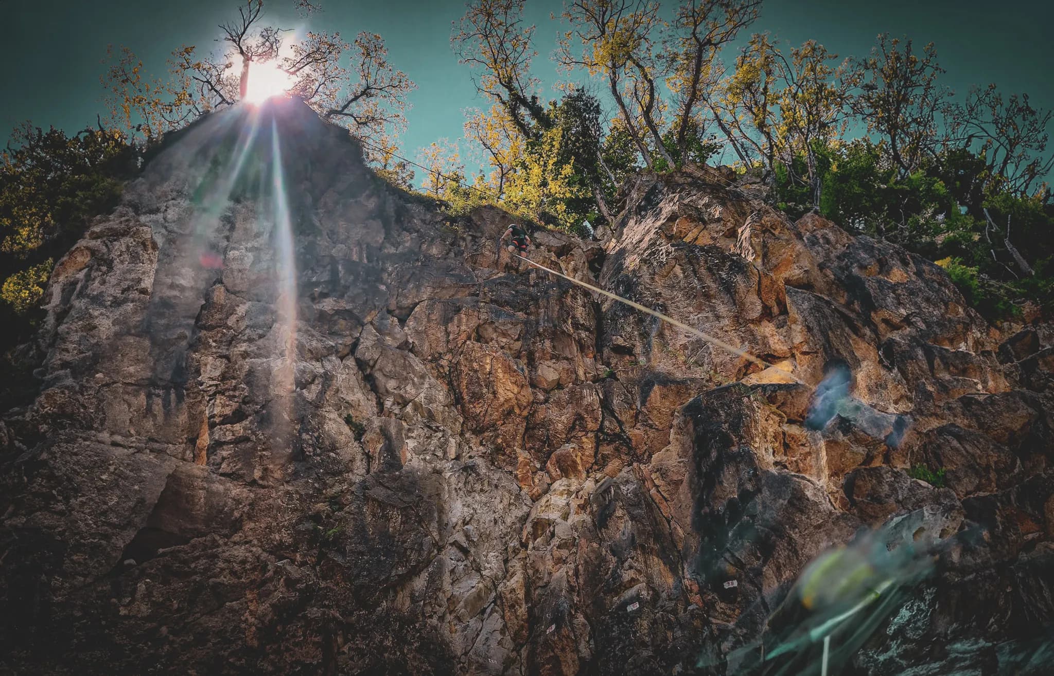 A climber suspended by a rope climbs a rock face, while the sun shines in the background, illuminating the scene with rays of light. The rock shows shades of brown and grey