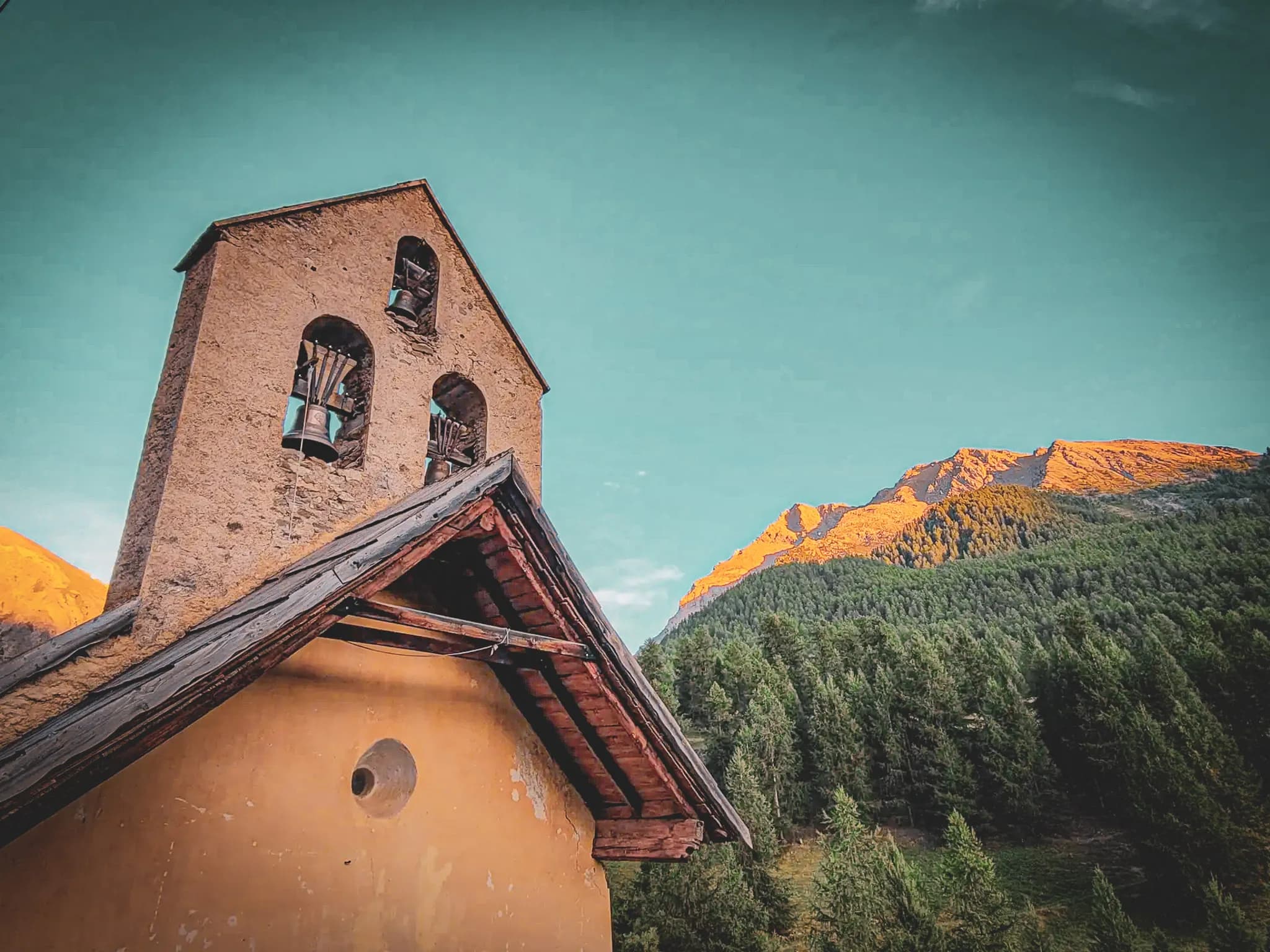 A stone bell tower overlooks a wooden hut with a sloping roof. Two bells can be seen through the windows in the bell tower. In the background, majestic mountains bordered by lush green forests.