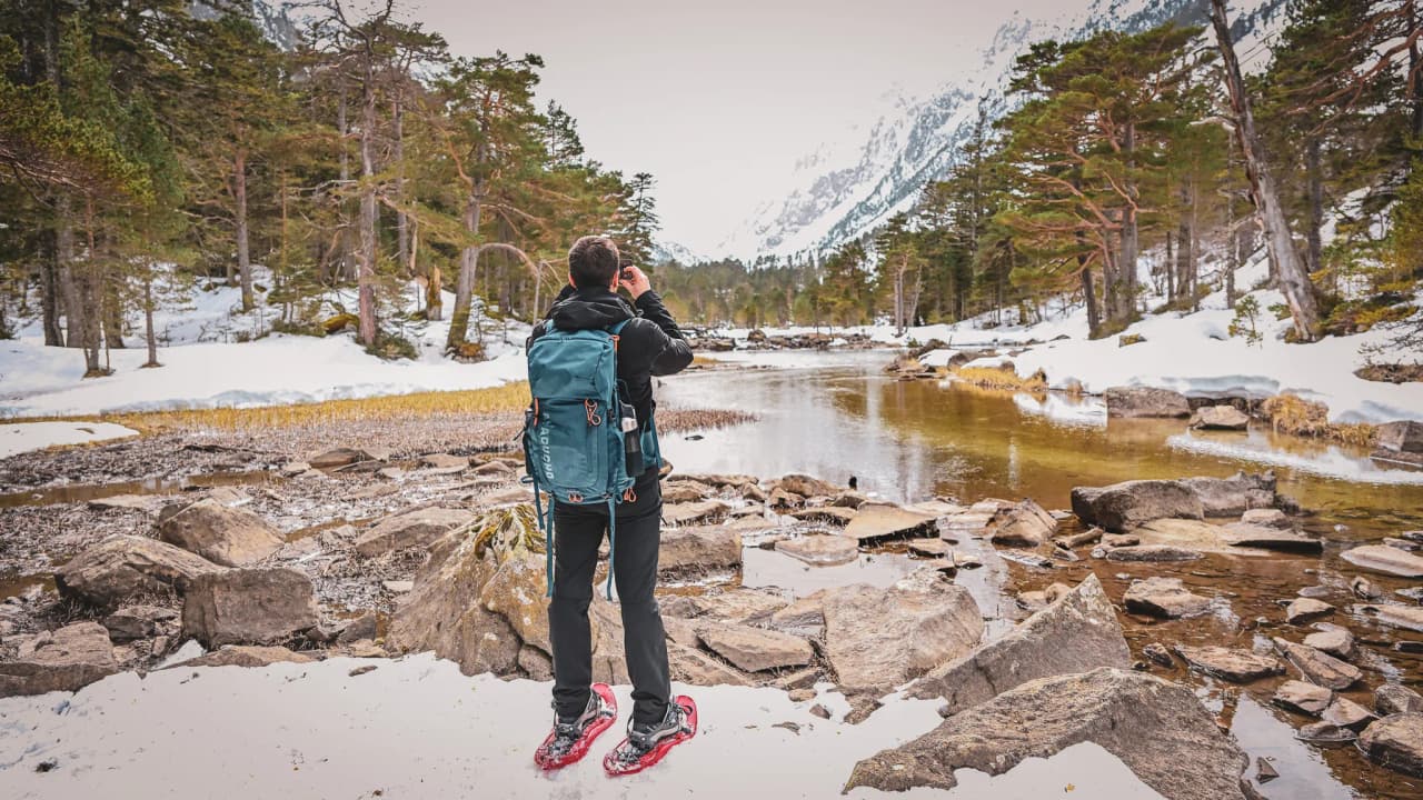 A snowshoe hiker admires a snow-covered mountain landscape beside a peaceful river.