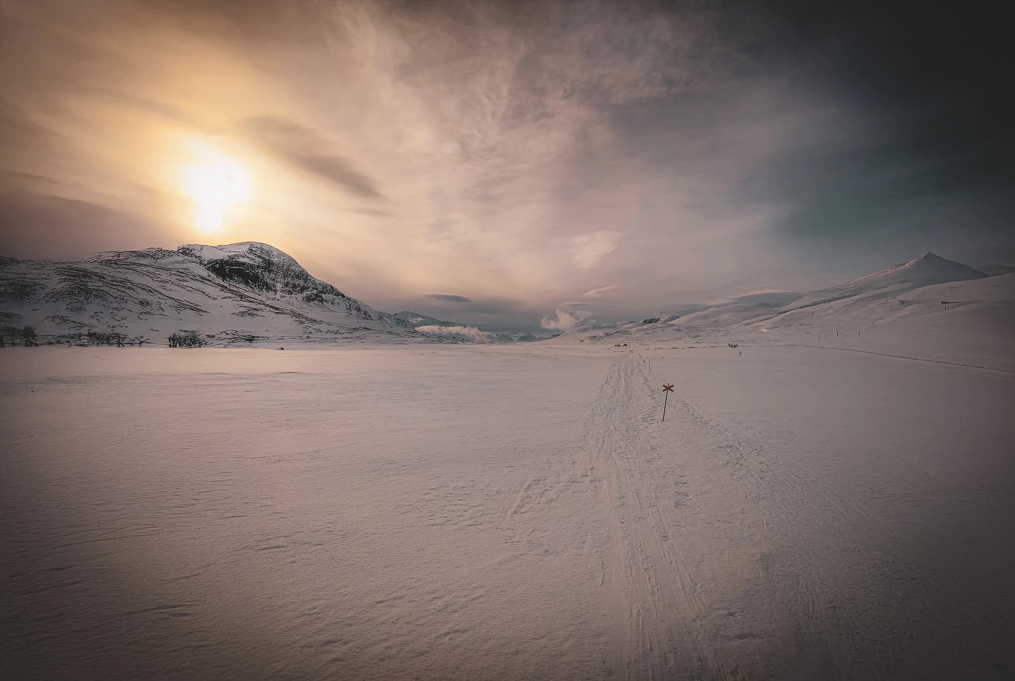 The snowy landscape of Kungsleden with mountains in the background. The sky has softly tinted clouds, while a sunrise or sunset illuminates the horizon with golden light.