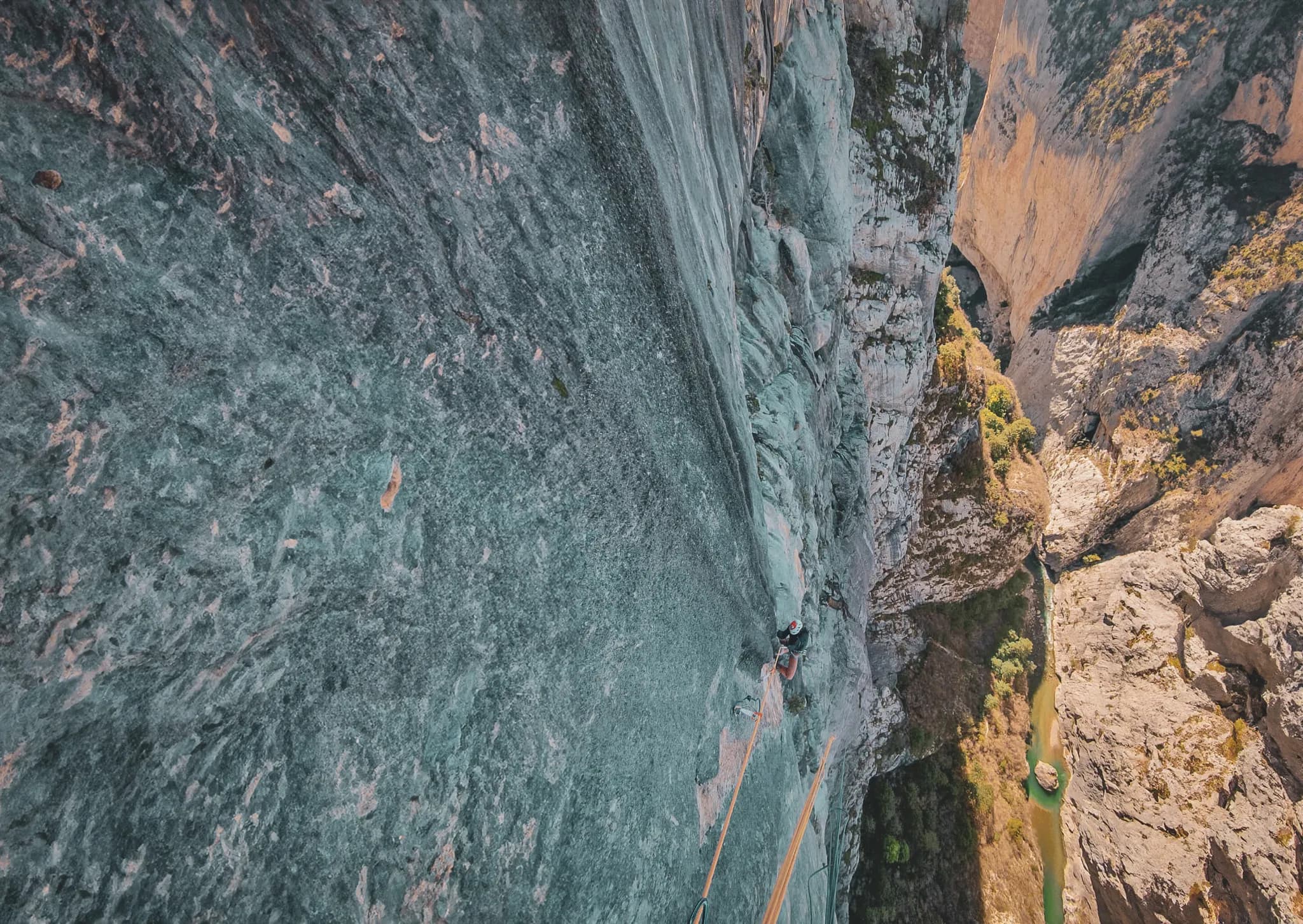 A climber ascends an imposing cliff, showing smooth, sheer rock faces. Below, a lush green valley with a stream runs through the landscape, while rock formations