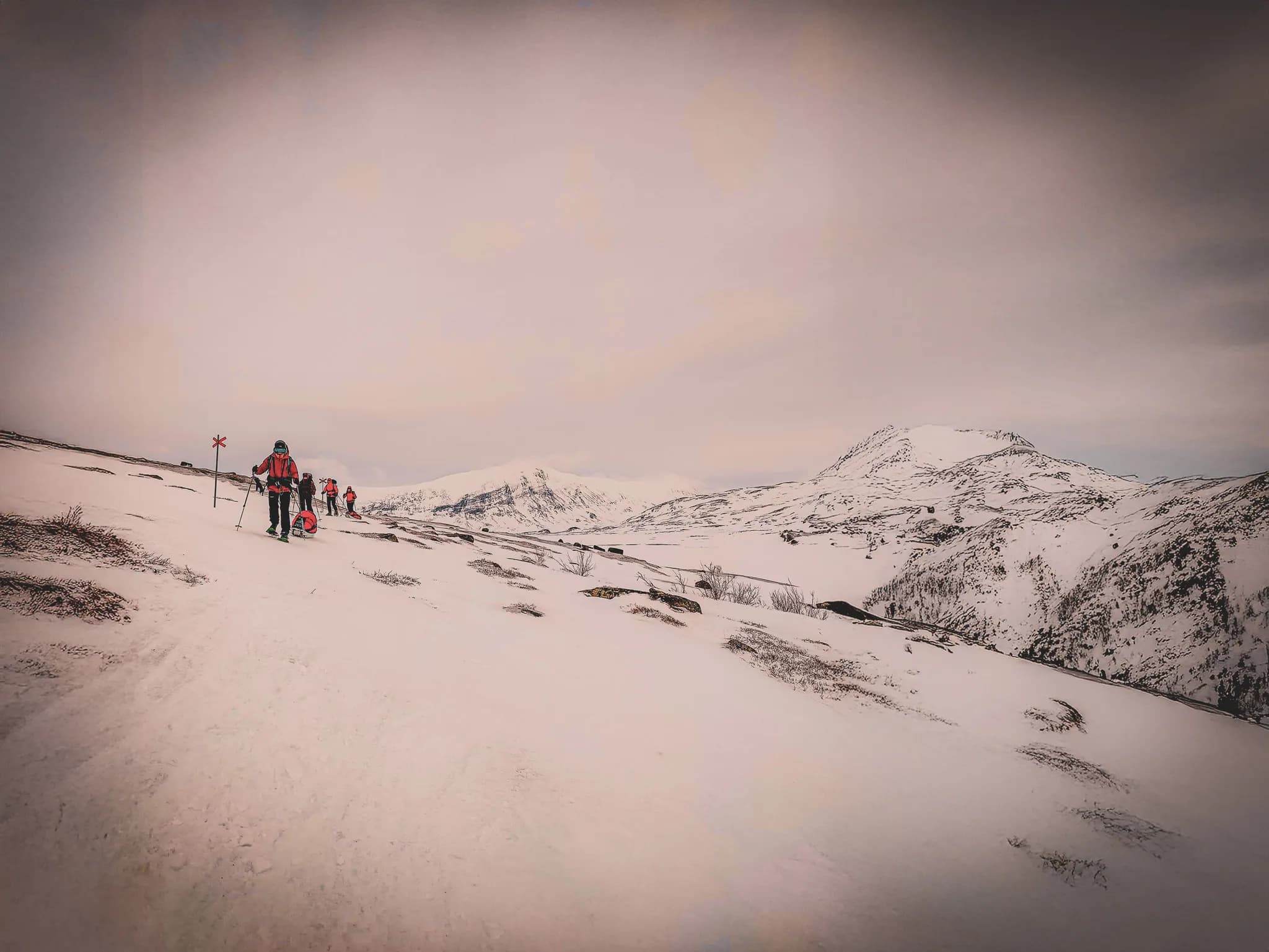 A group of mountain hikers make their way across snow-covered terrain, equipped with warm clothes and ski gear. In the distance, majestic peaks rise up under a slightly cloudy sky.
