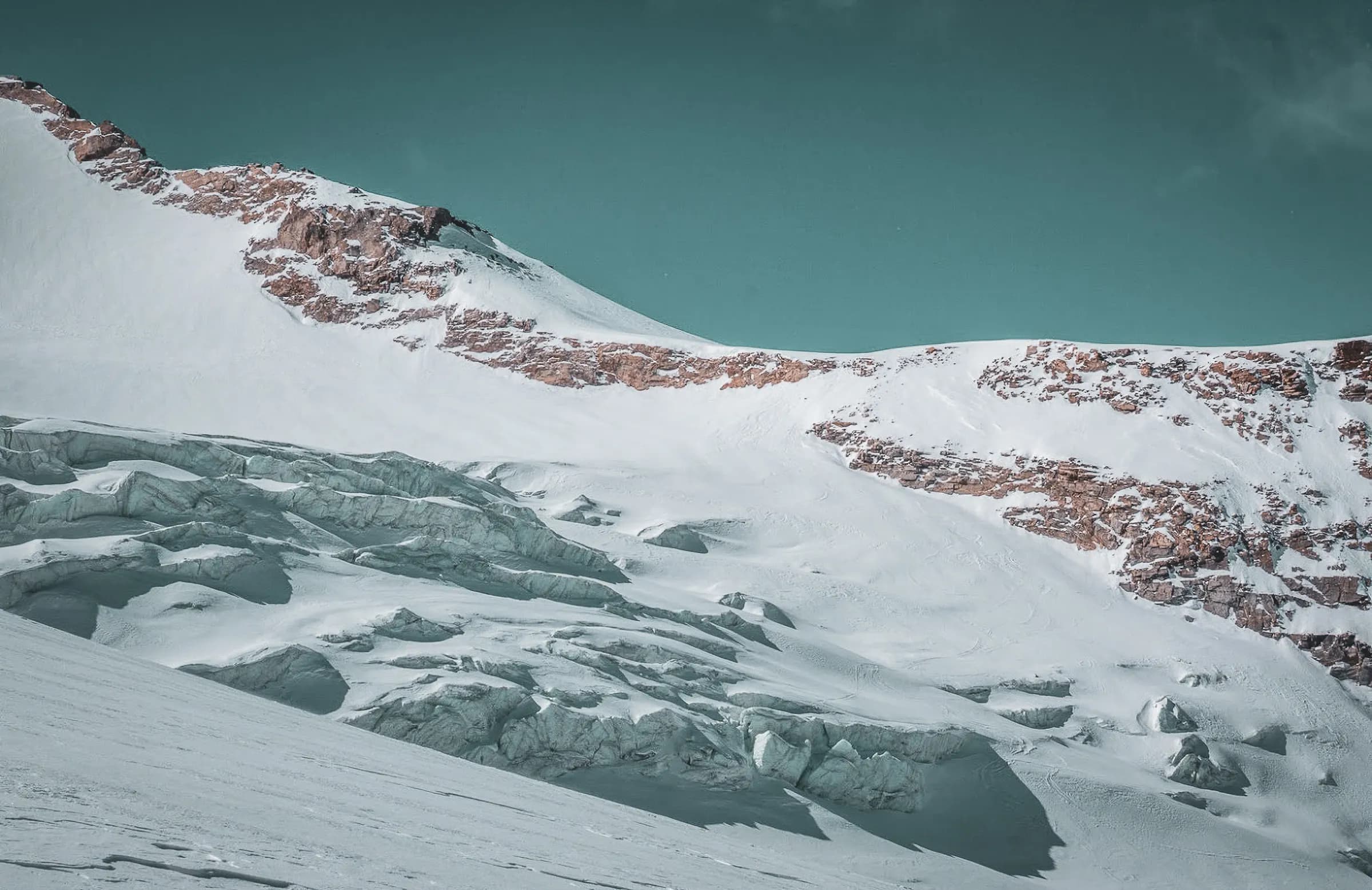 A mountainous landscape with snow-capped peaks and glaciers. In the background, steep rocky mountains stand out against a blue-green sky. In the foreground, glacial formations and snow recede into the distance.