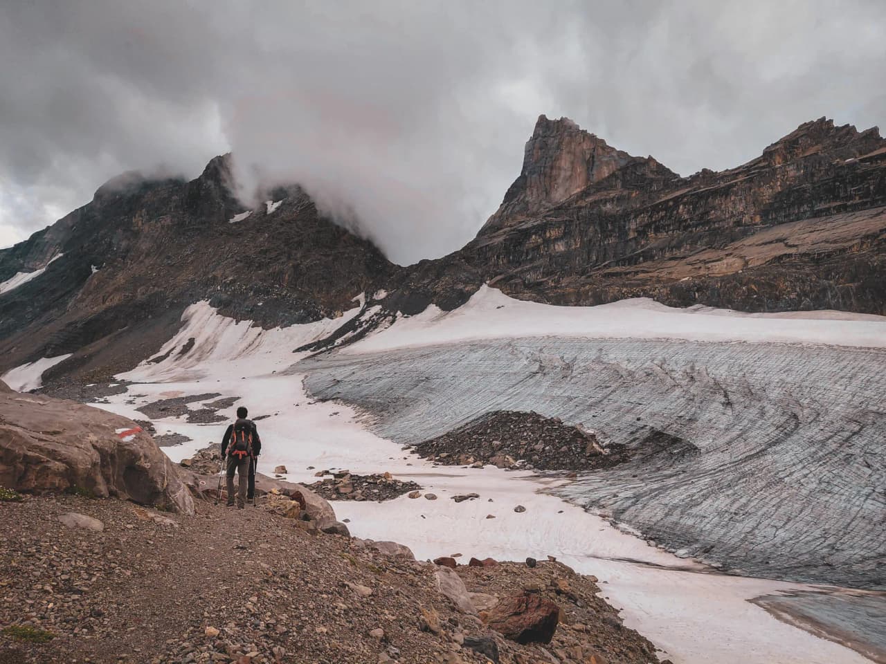A hiker contemplating a majestic glacier under a cloudy sky in the heart of the Alps.