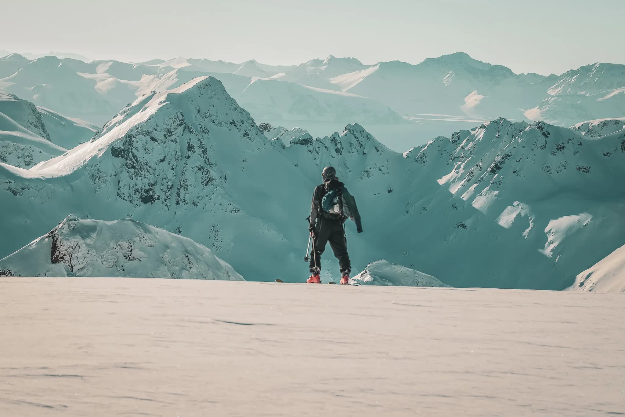 Un aventurier se tient sur un vaste terrain enneigé, face à une chaîne de montagnes majestueuses recouvertes de neige. Le ciel clair et lumineux accentue le paysage alpin, tandis que l'individu, équipé de skis