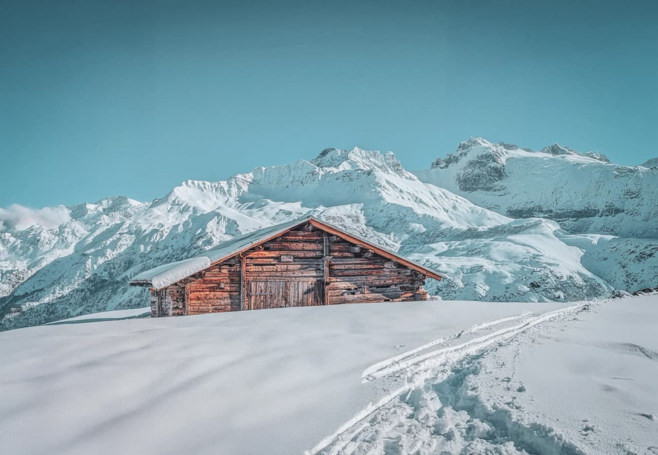 A wooden chalet stands in the middle of a vast snowfield, with majestic mountains in the background. The sky is a light blue, creating a striking contrast with the immaculate white of the snow. Visit