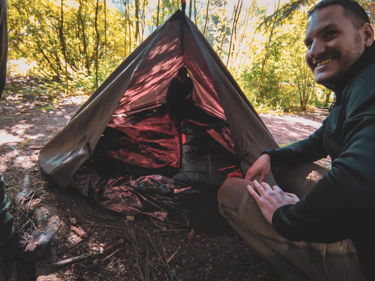Un participant souriant à un stage de survie, assis près d'une tente en pleine nature verdoyante.