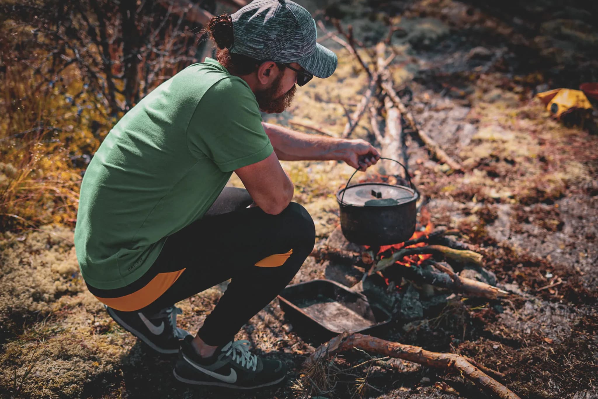 A man is crouched beside a campfire, holding a metal saucepan over the flames. He is wearing a green t-shirt and black sports trousers with orange accents. He has sunglasses and a case