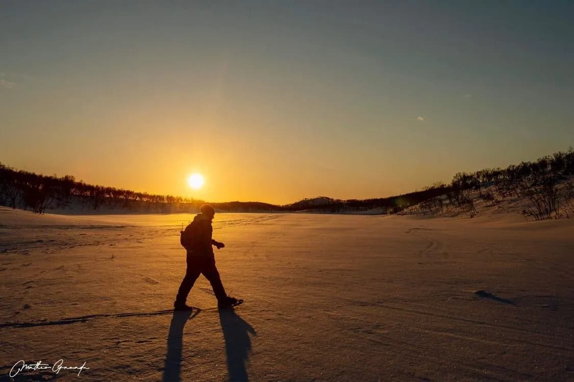 An adventurer walks alone across a vast snowfield, illuminated by the setting sun. The person's silhouette is silhouetted against a sky of warm orange and blue, while wooded hills rise up to the horizon.