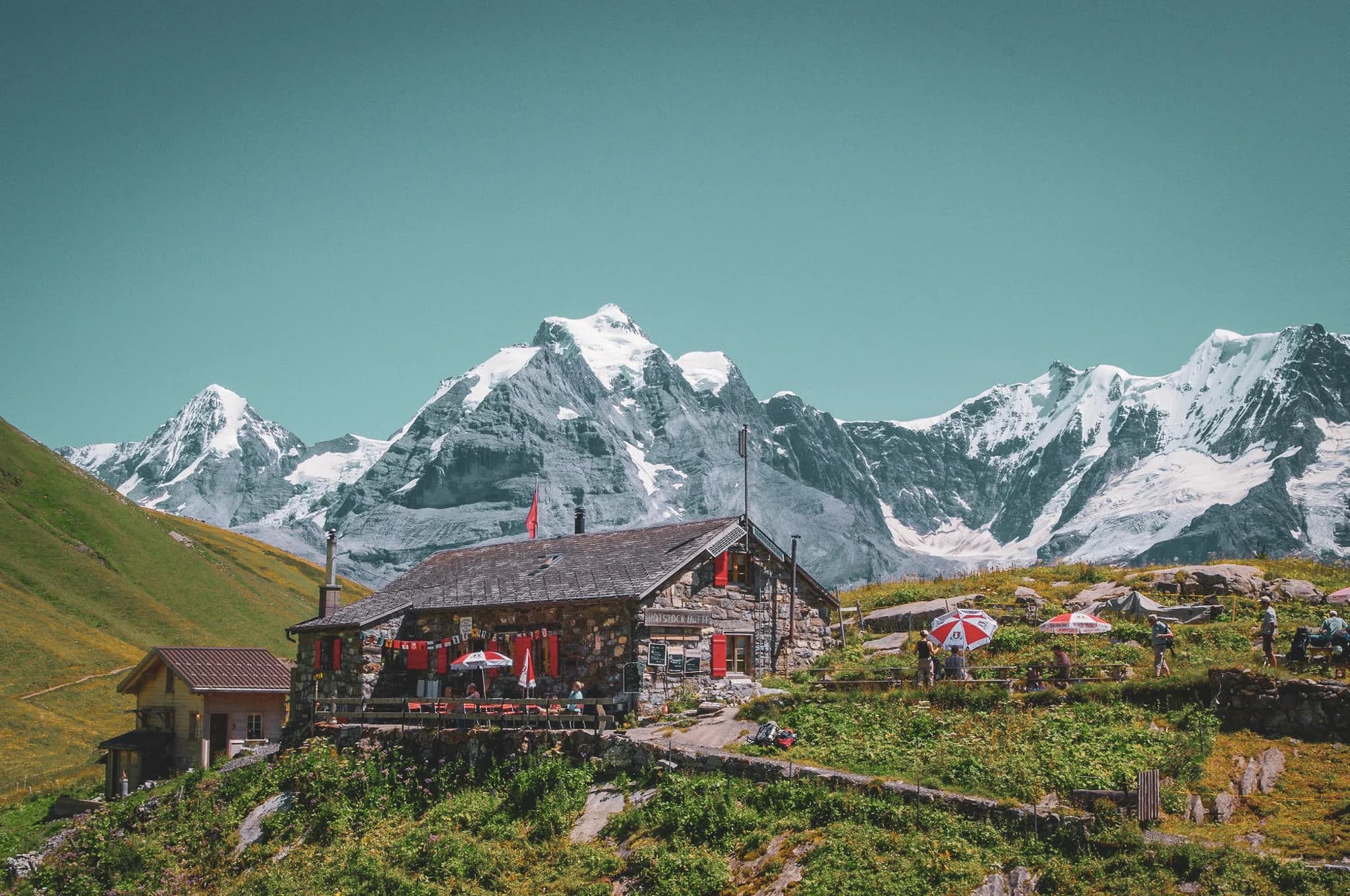 A stone chalet with a tiled roof is set in a mountain setting. Colourful flags and parasols decorate the terrace, where people gather to enjoy the view.