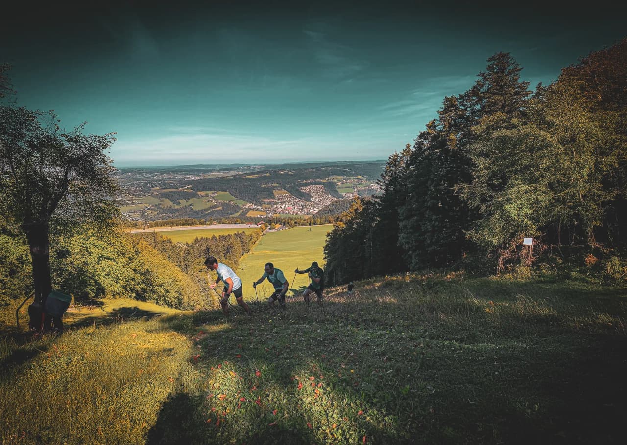 Een groep wandelaars beklimt een groene heuvel met een panoramisch uitzicht op de Jura.