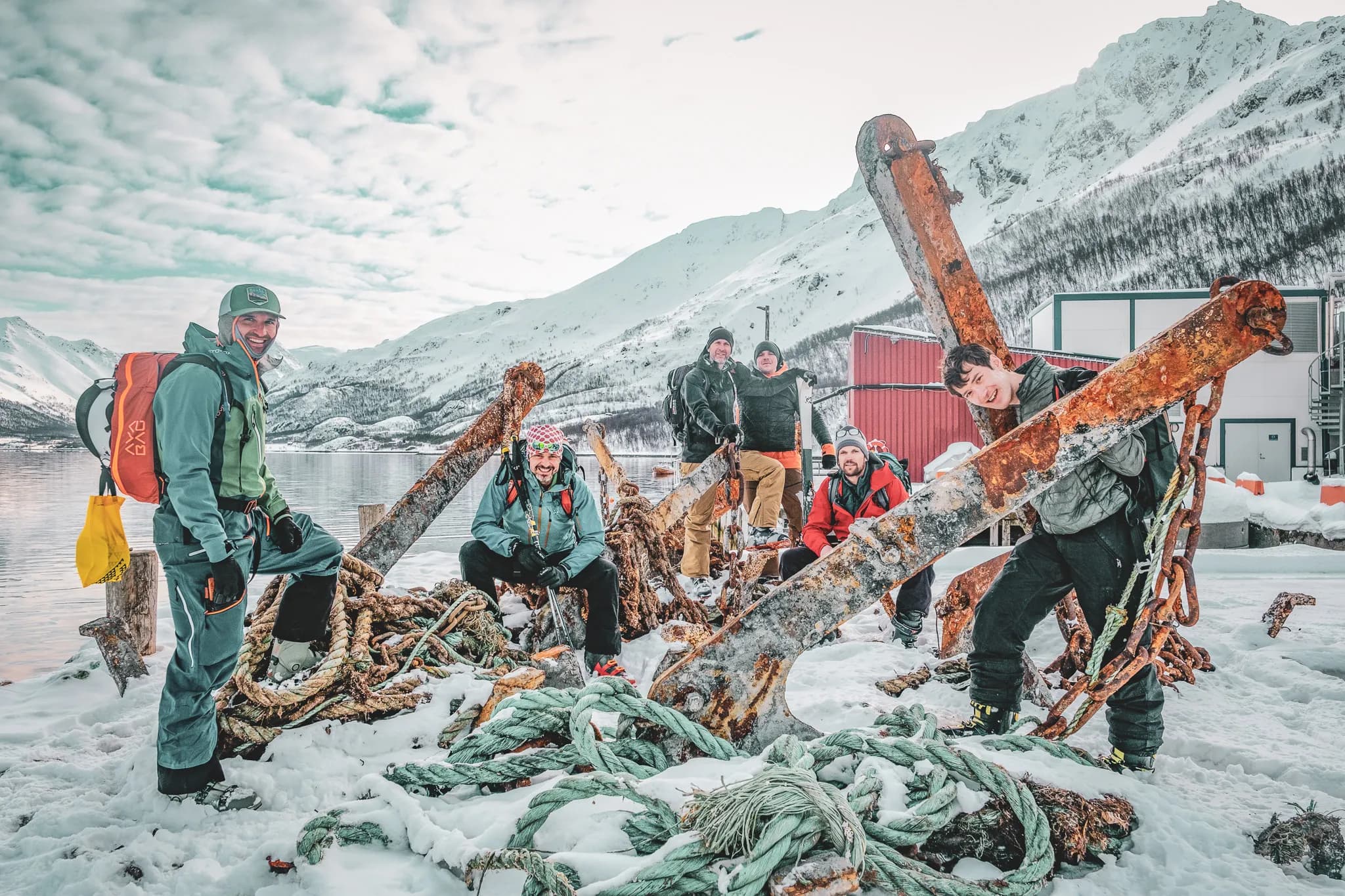 Un groupe de six personnes posent joyeusement autour d'une énorme ancre rouillée, recouverte de neige et entourée de câbles marins. Le paysage est composé de montagnes enneigées et d'une étendue d'eau calme.