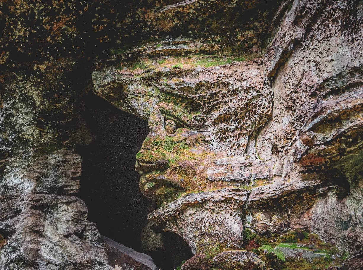 A natural sculpture in rock shows a stylised human face, with distinct features such as eyes and a mouth. The surface of the rock is textured, with cracks and irregular patterns. Touches