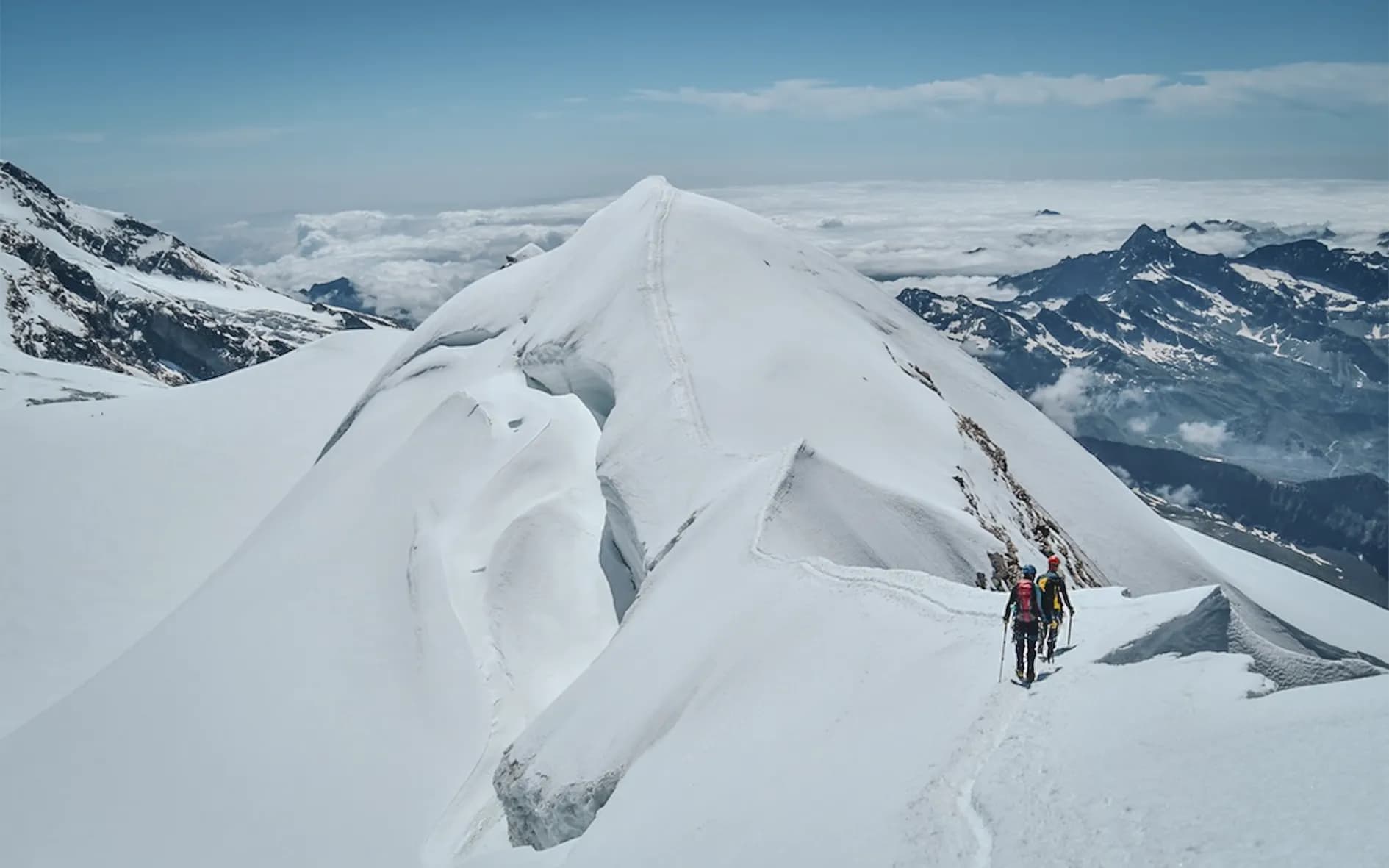 A majestic snow-capped peak stands out against an Alpine landscape of white slopes and steep ridges. In the foreground, a group of hikers are making their way up a sent