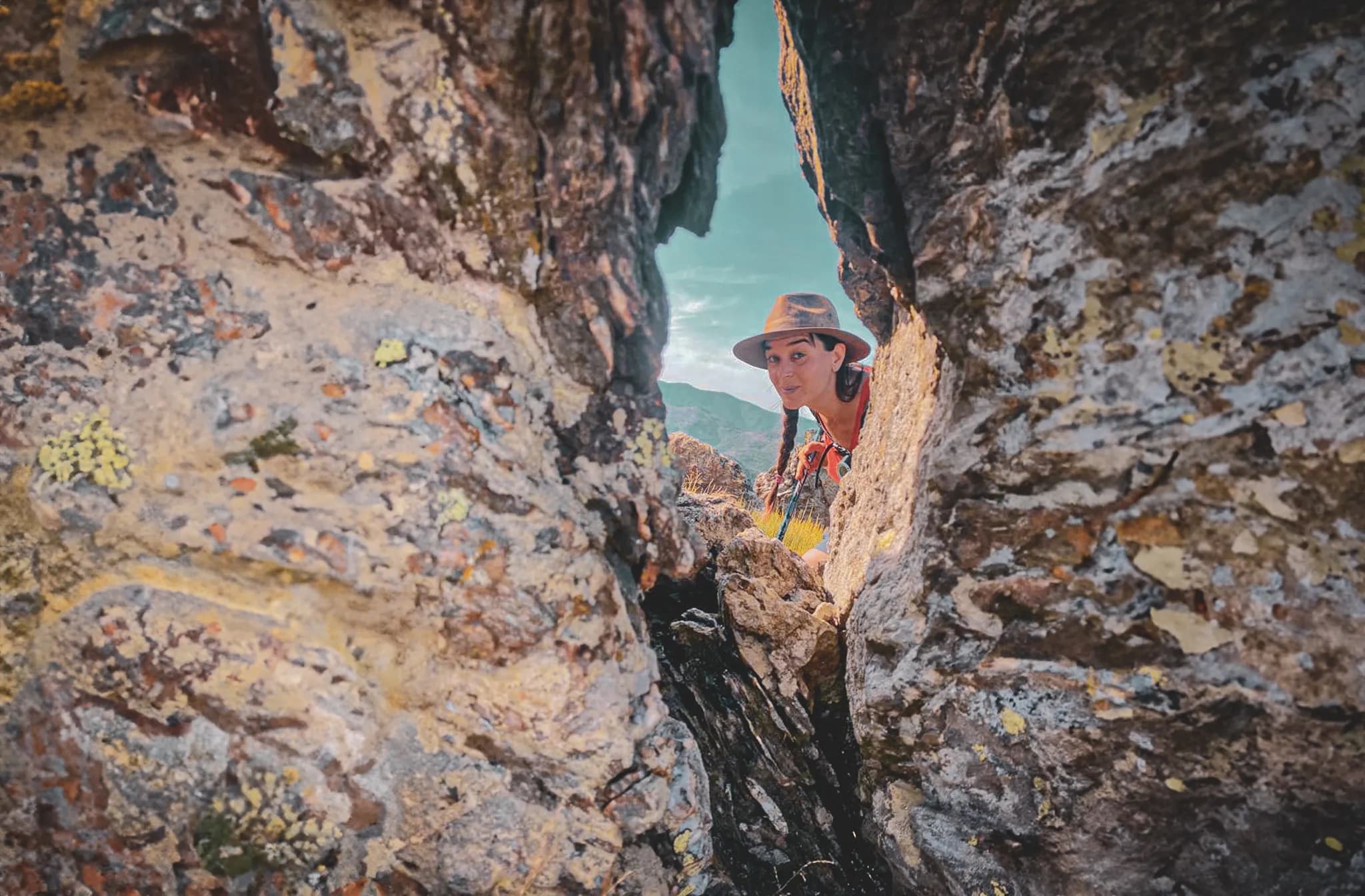 A rocky landscape reveals a smiling woman emerging from a hollow between two large stones. She is wearing a hat and a colourful outdoor outfit, with plaits falling over her shoulders. The