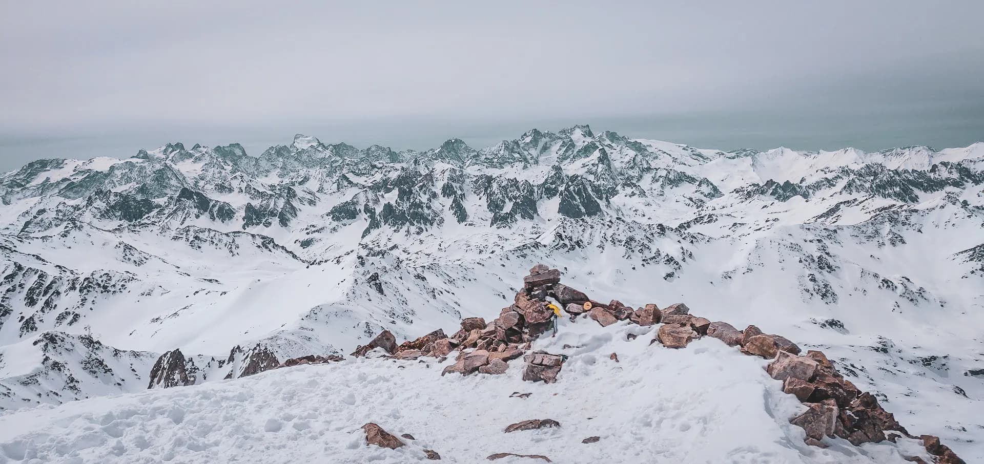 Un paysage montagneux majestueux, recouvert de neige, avec des sommets escarpés se dressant à l'horizon. À l'avant-plan, un petit monticule de pierres, probablement un cairn, se trouve sur un sol