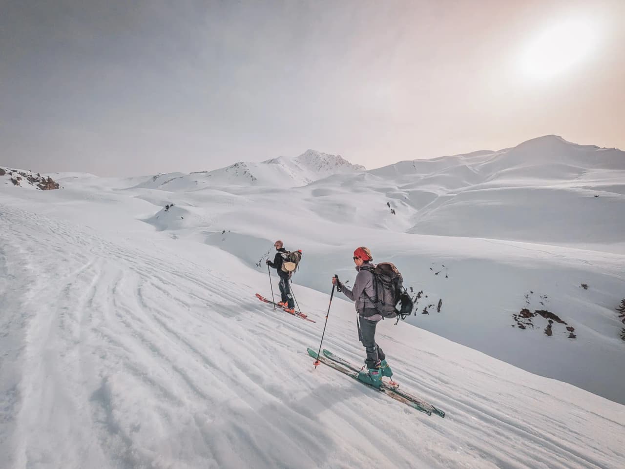 Two cross-country skiers advance across a snow-covered landscape, with majestic mountains in the background, under a clear sky.