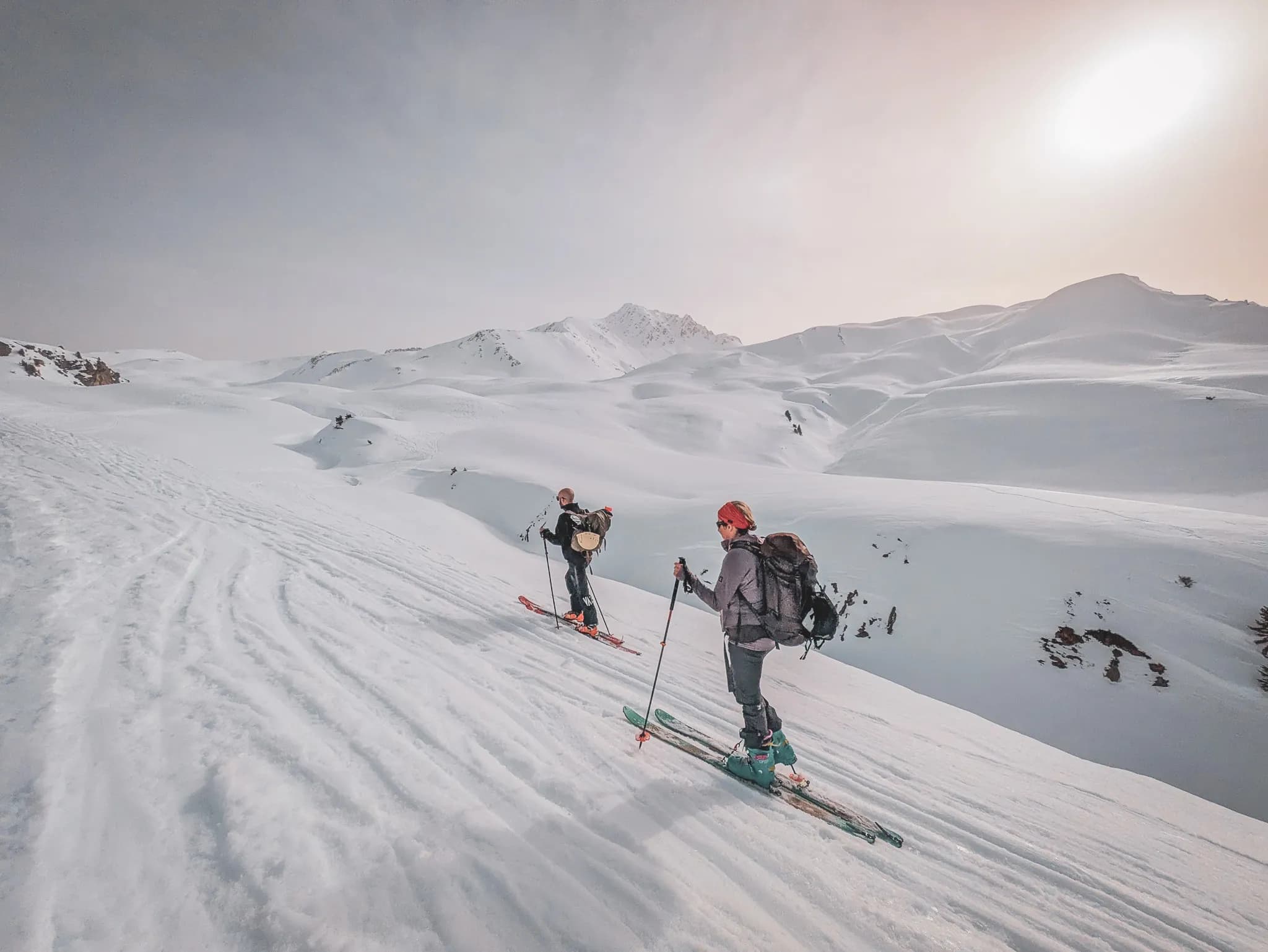 Two cross-country skiers advance across a snow-covered landscape, with majestic mountains in the background, under a clear sky.