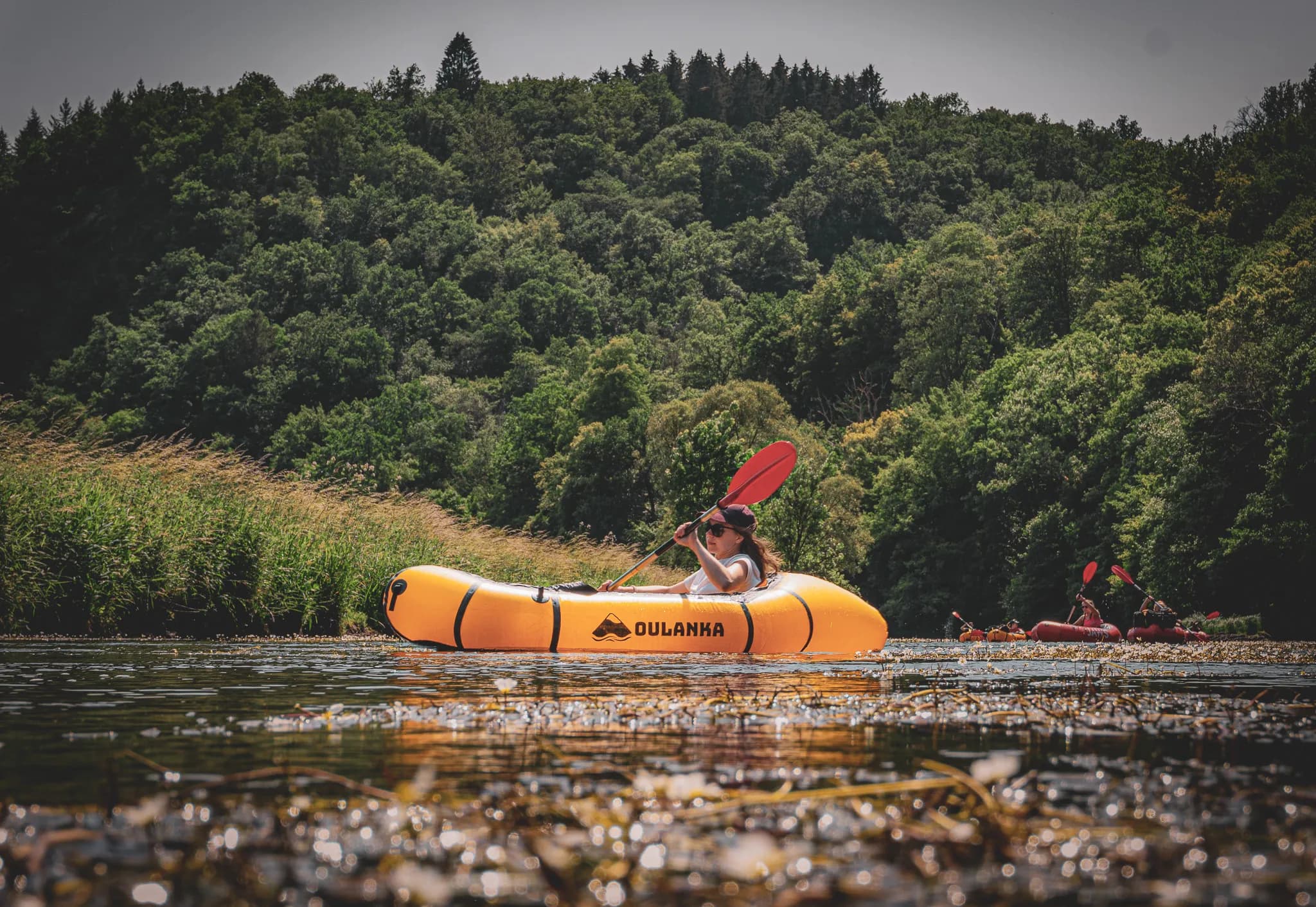 Een oranje kajak peddelt op een kalme rivier, omgeven door weelderig groen van heuvels en bomen. Eén persoon peddelt in de kajak en draagt een pet en een zonnebril. Op de