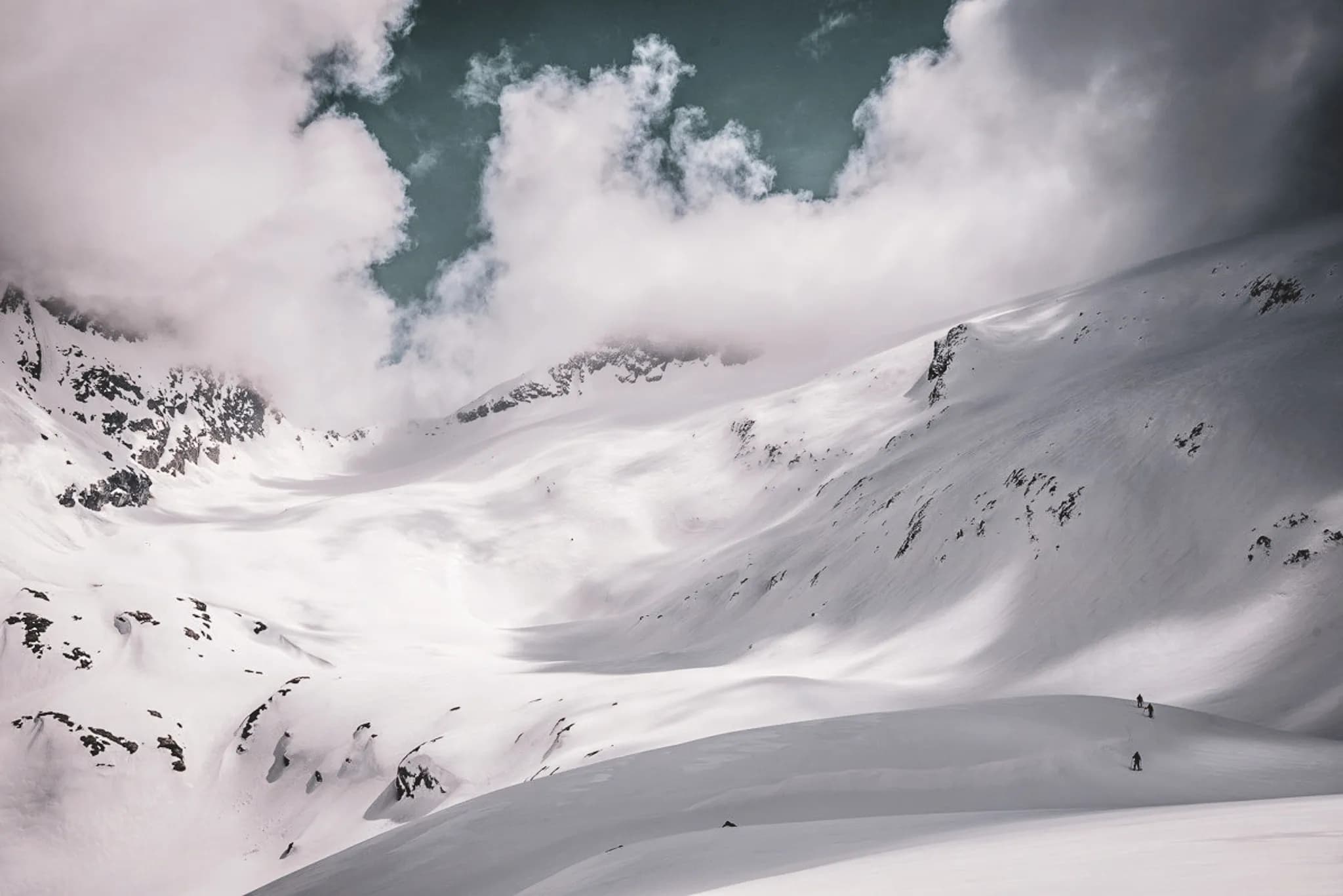 A snow-covered mountain landscape, with steep ridges looming on the horizon. Thick, dark clouds float above, surrounding the peaks. In the foreground, two human figures