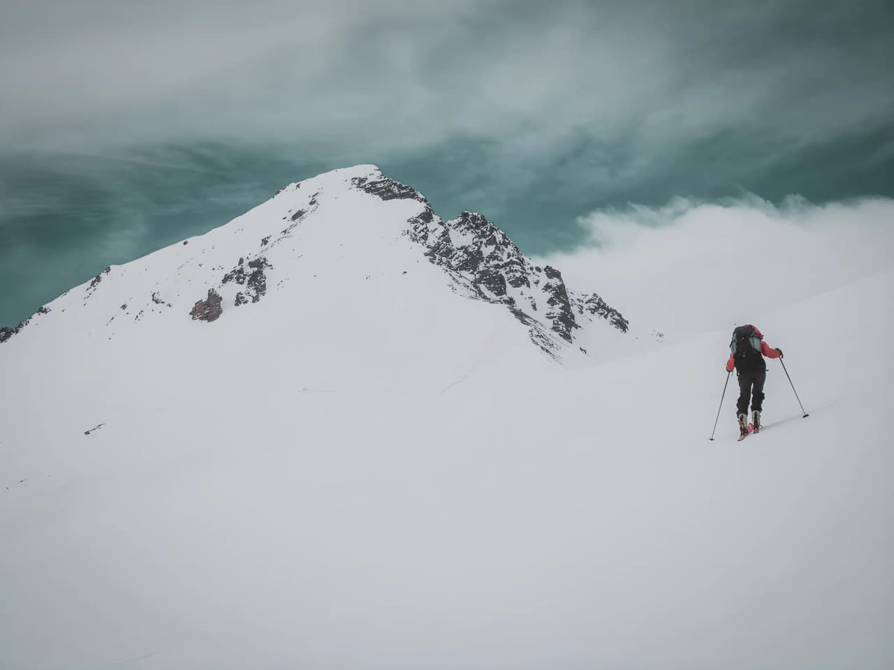 Un skieur solitaire grimpe une montagne enneigée sous un ciel dramatique, invitant à l'aventure.