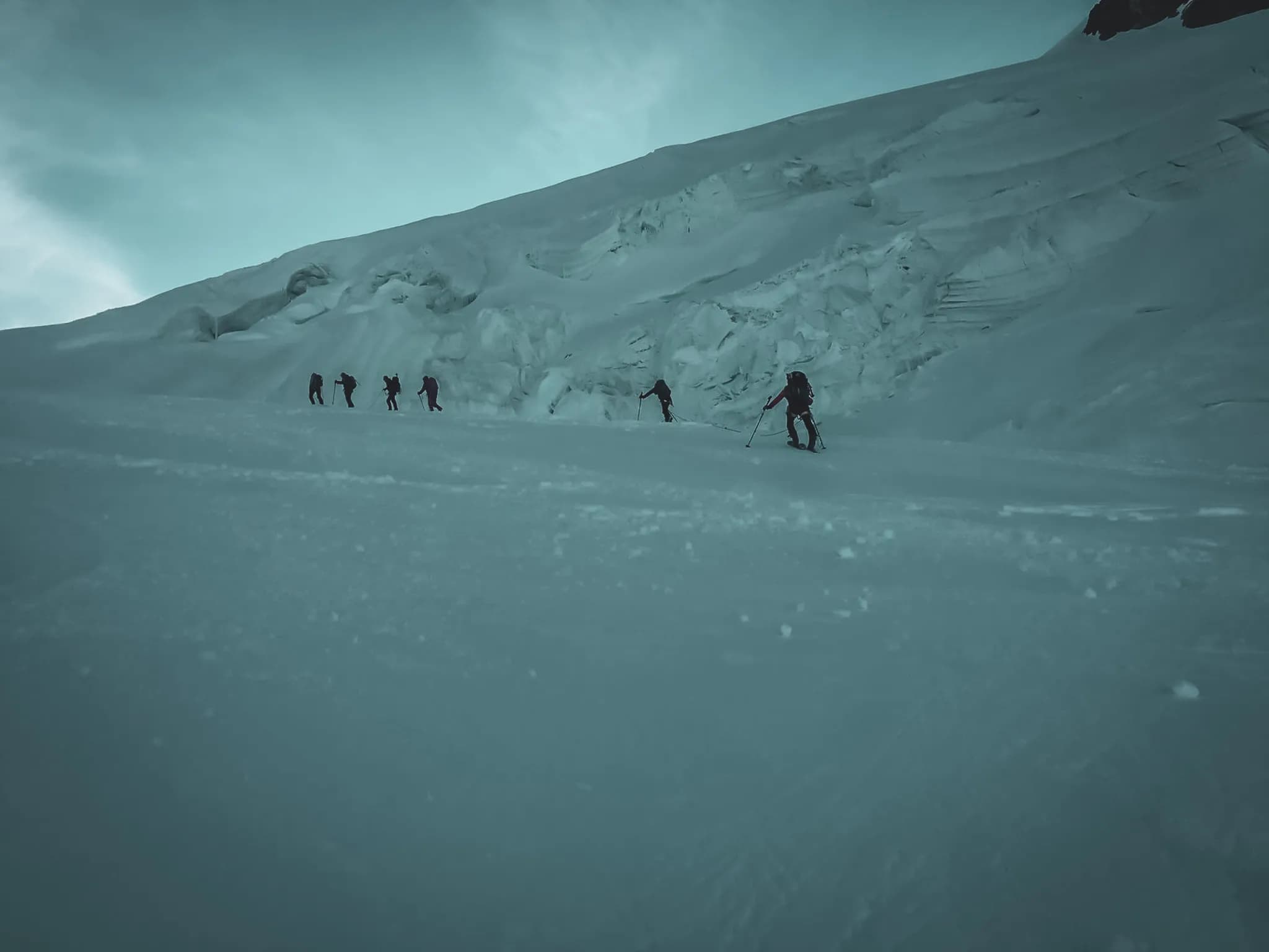 Een groep skiërs daalt af van een sneeuwhelling, met een gletsjerlandschap op de achtergrond. Het licht is zacht en creëert een serene sfeer. De silhouetten van de skiërs zijn zichtbaar, ze bewegen weg in de richting van de gletsjerformaties.