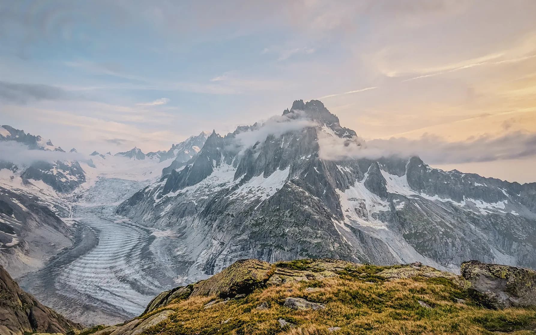 A spectacular mountain landscape with high, snow-covered peaks and clouds settling over the summits. Below, a glacier winds its way through a valley, showing streaks on its surface. The light