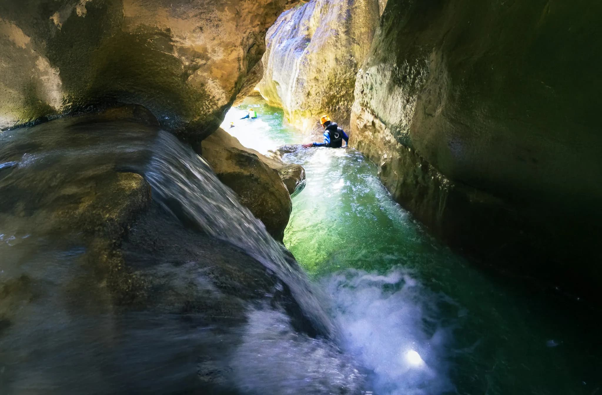 A narrow canyon with rock walls surrounding a river of turquoise water. Drops of water trickle over the rocks, creating a small stream. An adventurer in wetsuit and helmet