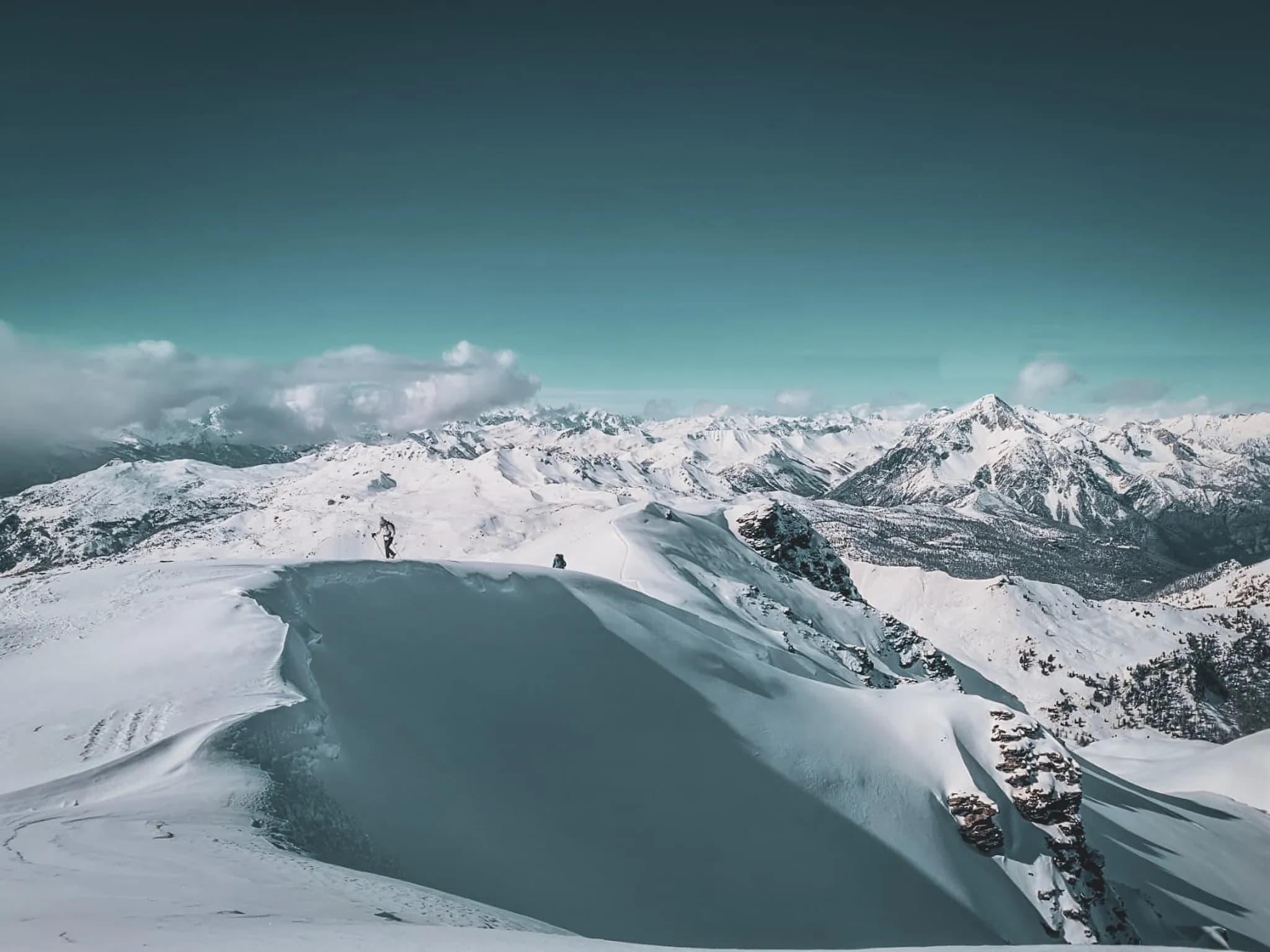 A snow-covered mountain landscape, with majestic peaks rising in the background. Two people, equipped with hiking gear, stand in the foreground. One of them is carrying a rucksack and